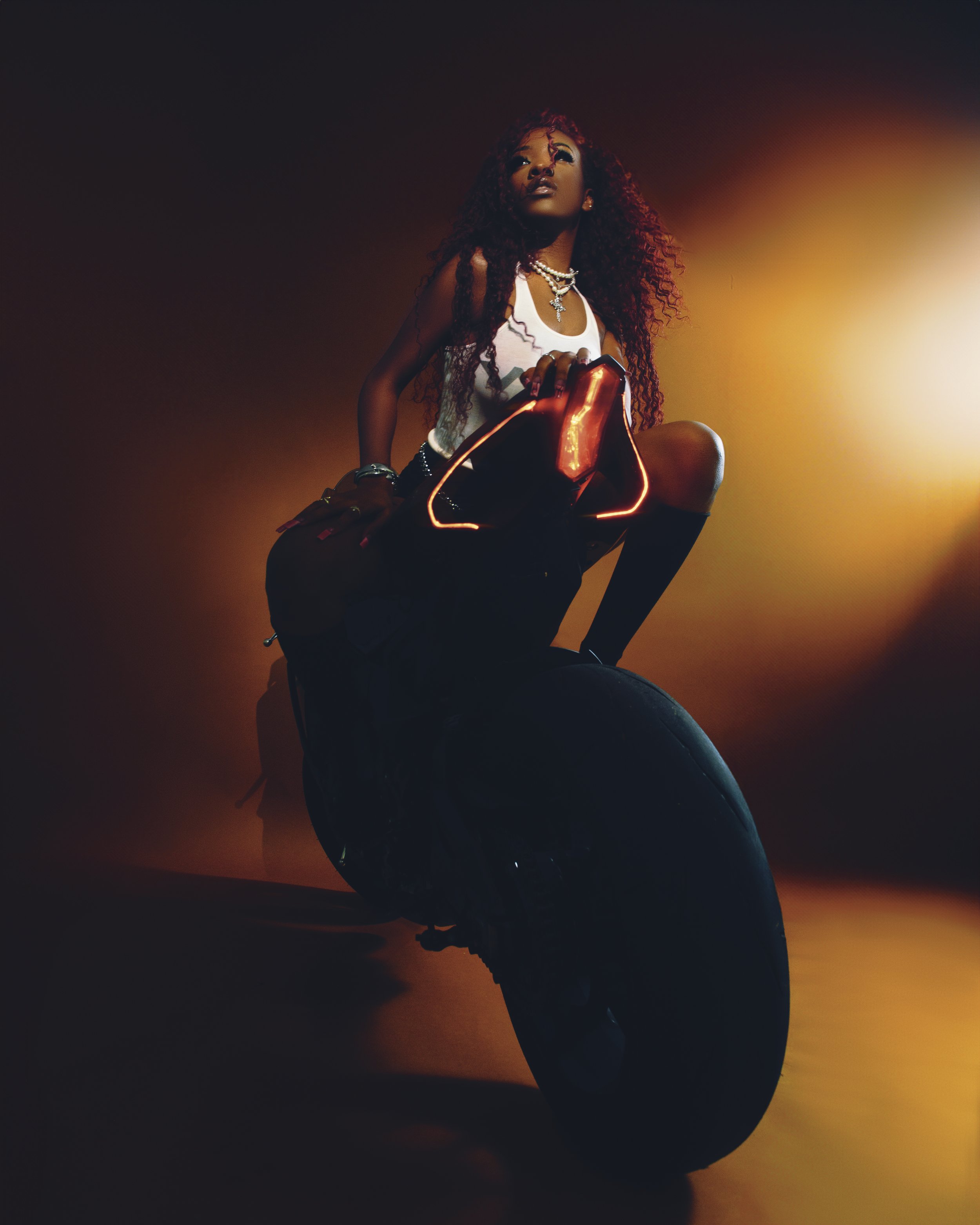 Stylish woman with long curly hair sitting in dramatic orange and red lighting, wearing layered necklaces and holding a glowing red handbag.
