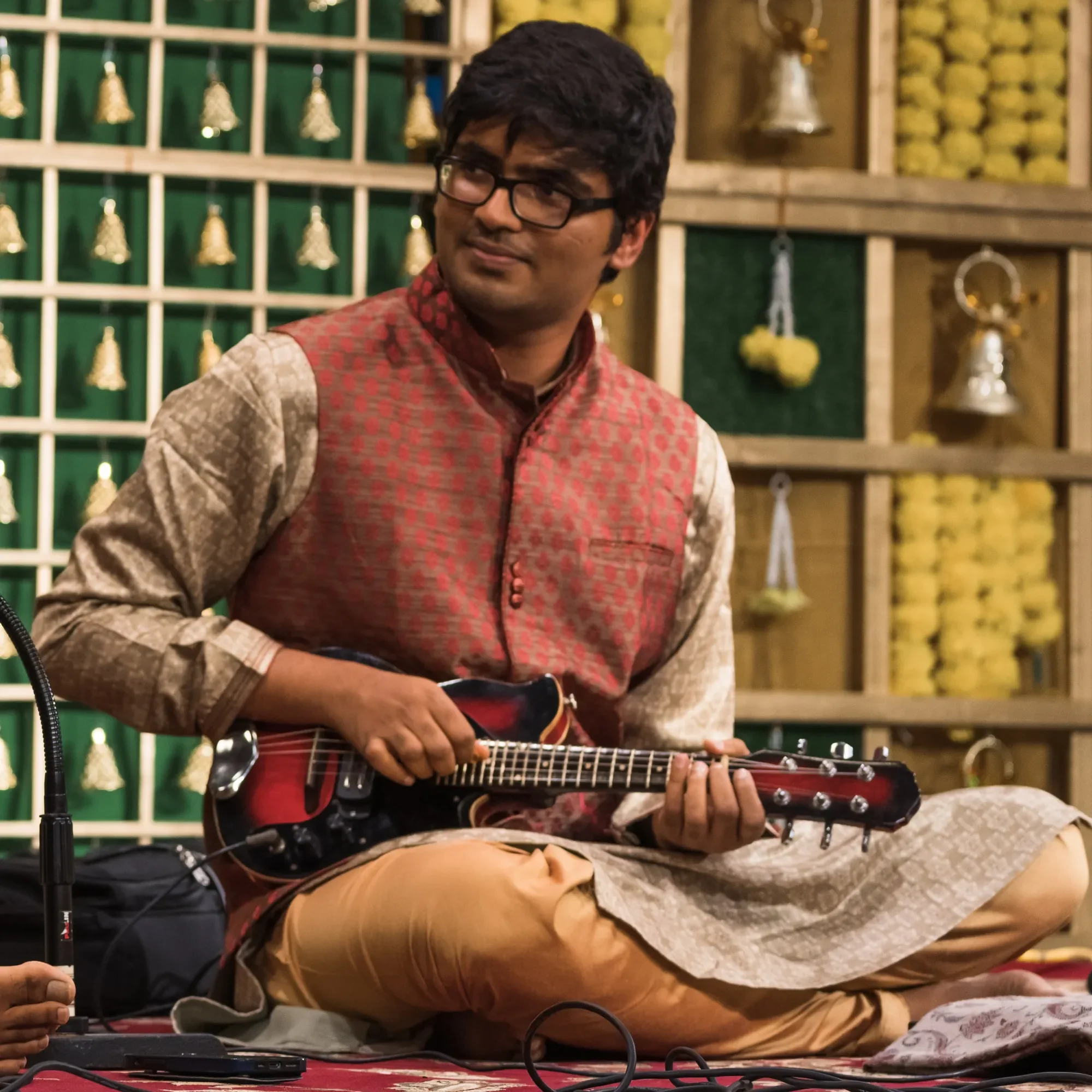 A man wearing glasses and traditional Indian clothing playing an electric mandolin on a stage decorated with bells and yellow flowers.