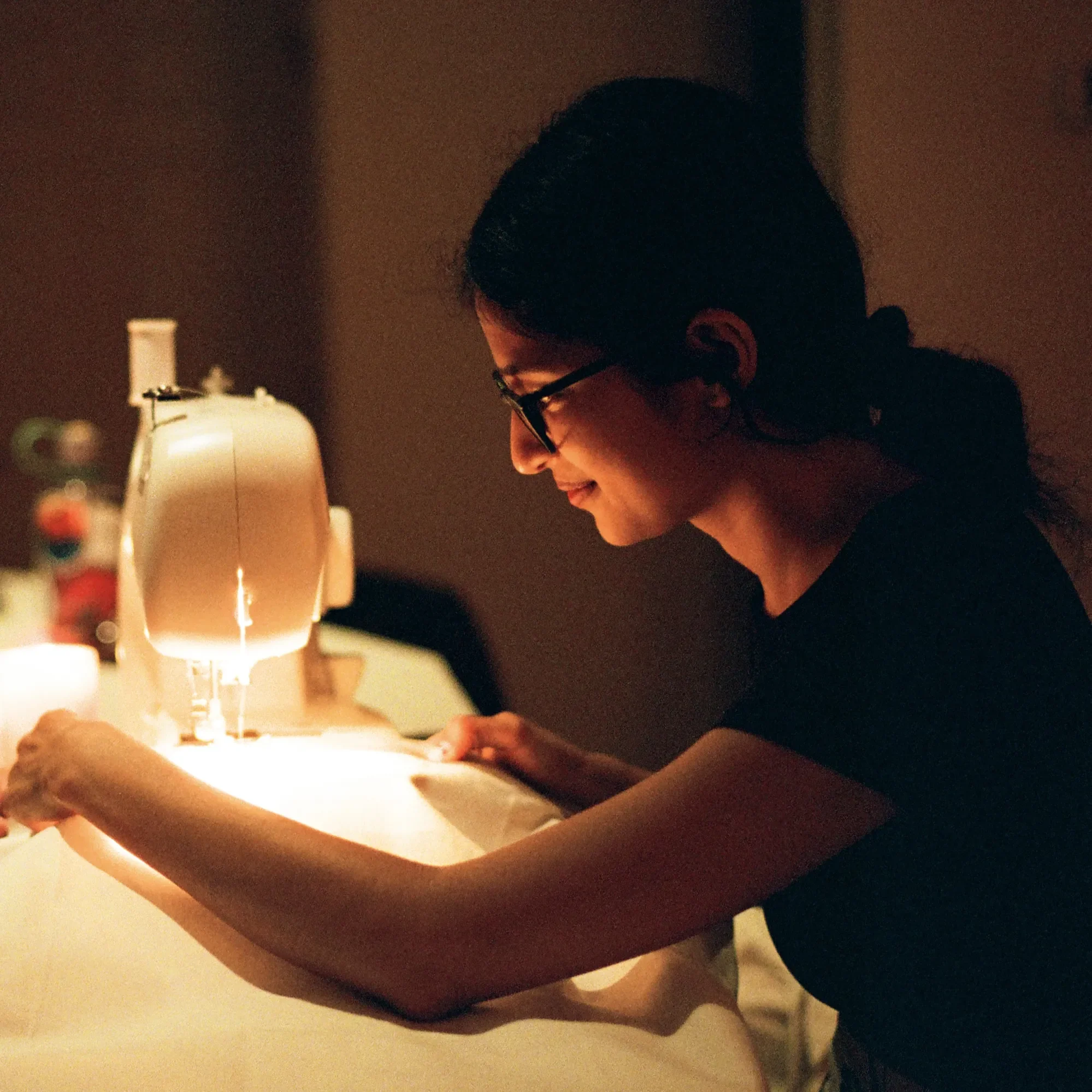 A woman in glasses using a sewing machine in a dimly lit room.
