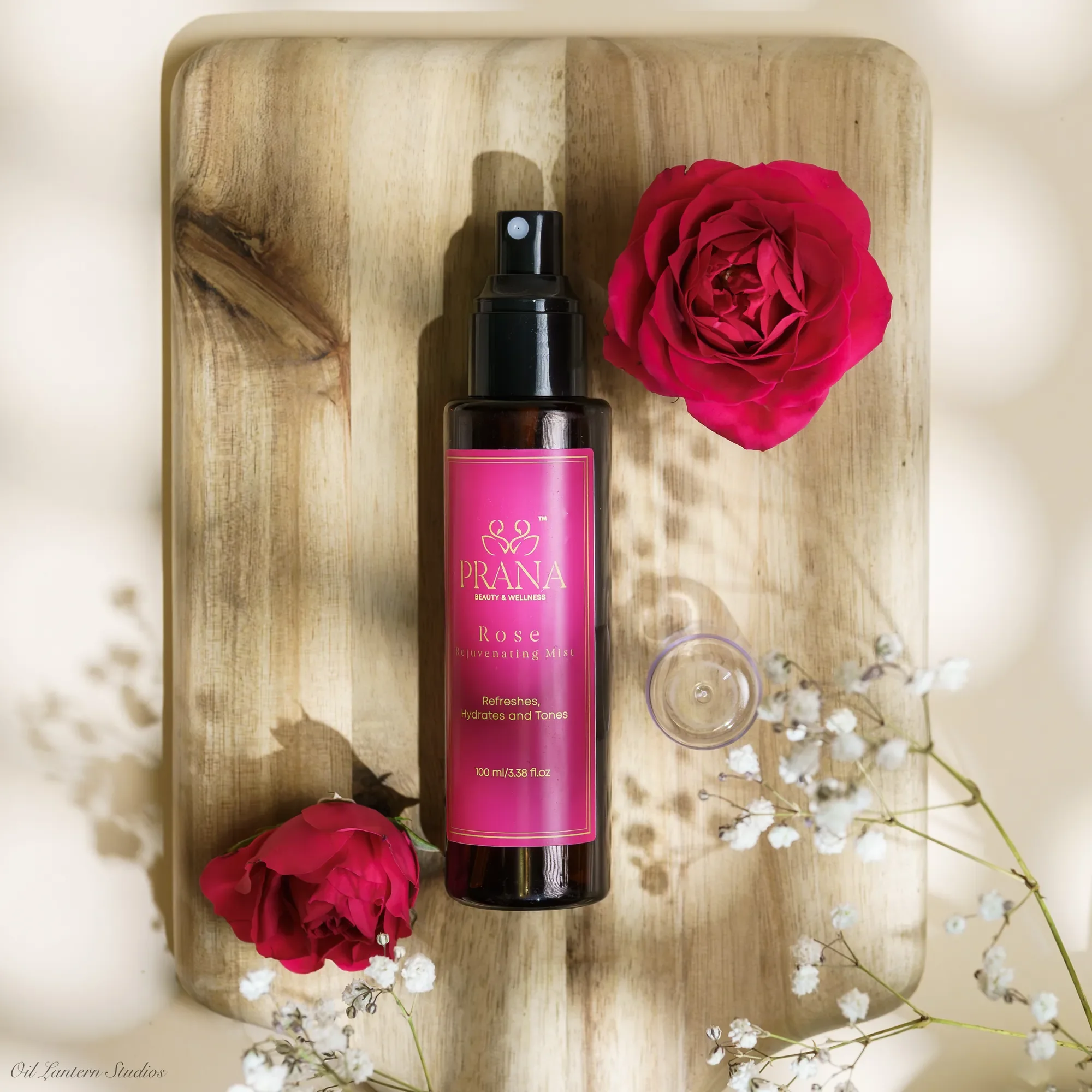 A pink bottle of Prana Rose Rejuvenating Mist on a wooden tray, accompanied by two pink roses and white baby's breath flowers.