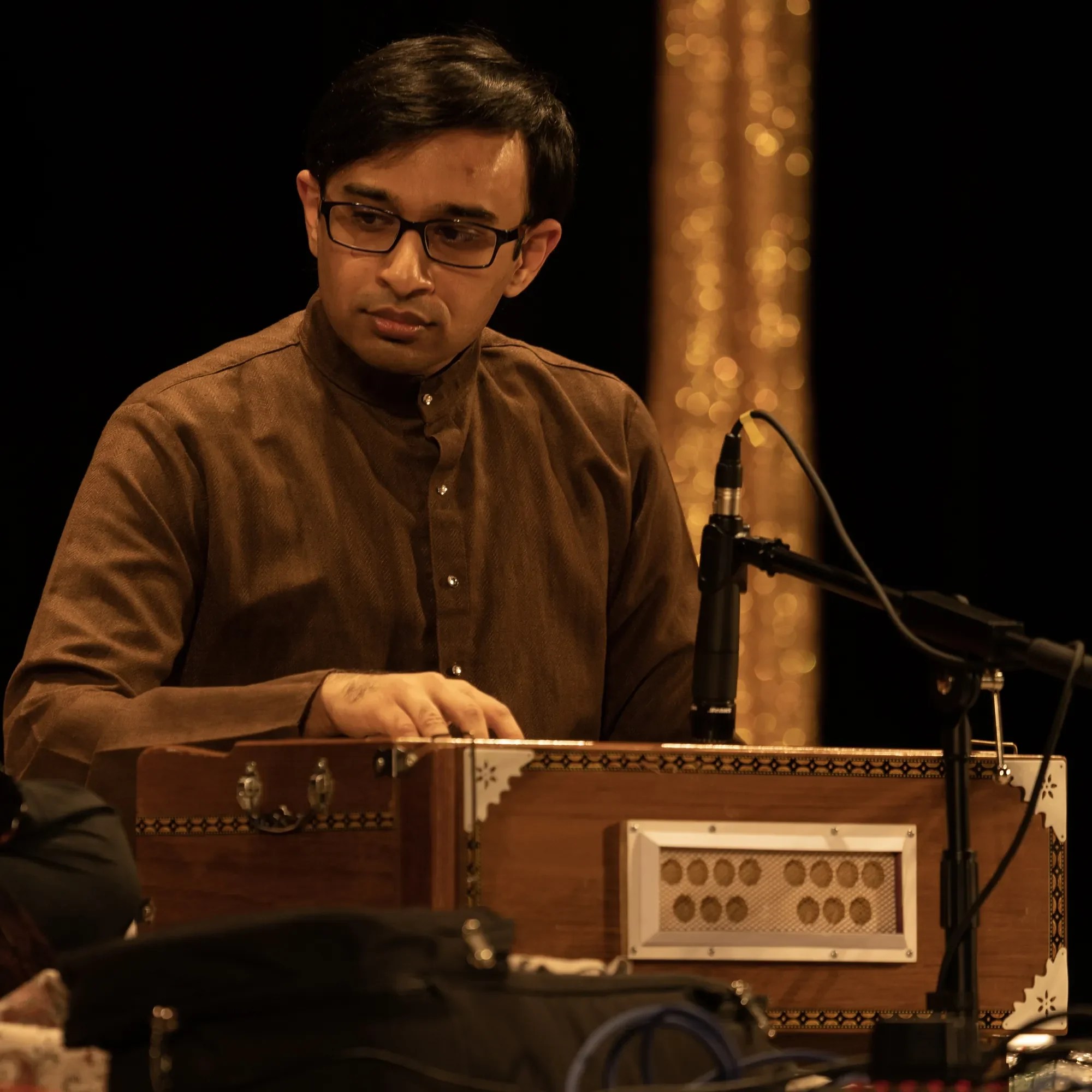 A man with glasses playing a harmonium on stage, wearing a brown kurta, with a microphone set up nearby, and a decorative gold background.
