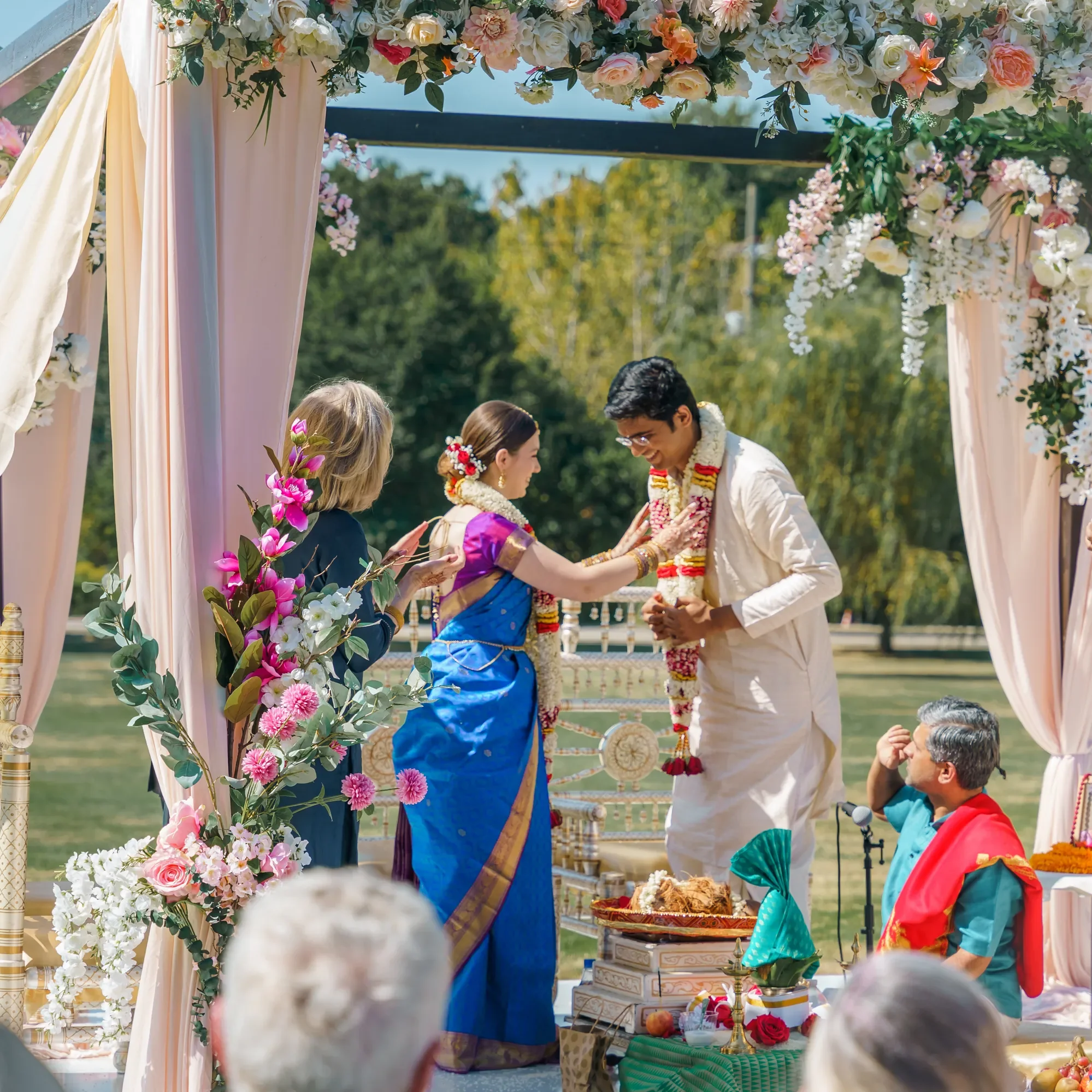 A traditional outdoor Indian wedding ceremony with a couple exchanging garlands under a floral decorated canopy, surrounded by family and friends.