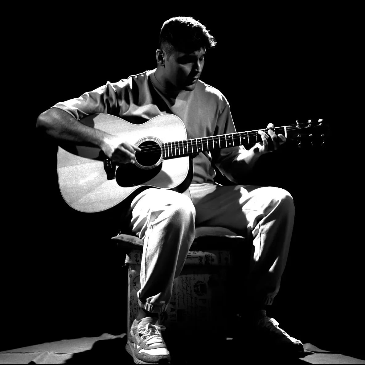 A young man playing an acoustic guitar on a dark stage, sitting on a decorated stool, with dramatic lighting highlighting his face and the guitar.