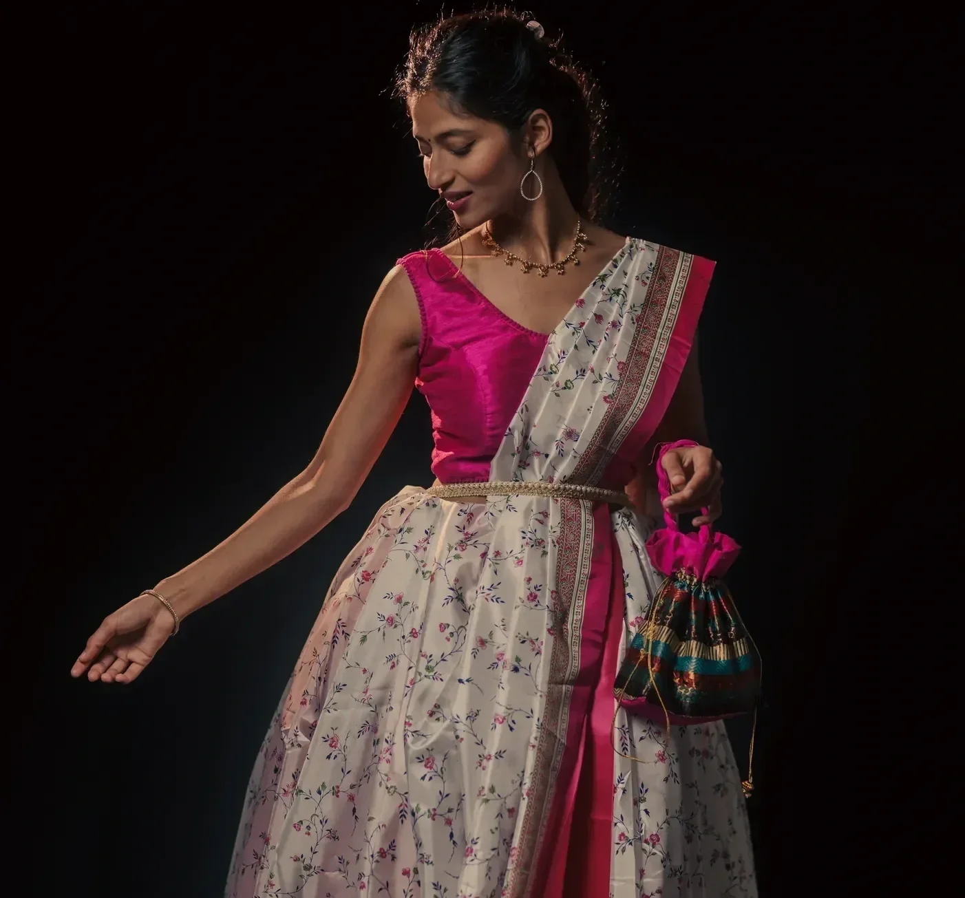 A woman in traditional Indian attire, wearing a pink blouse and cream-colored lehenga with floral patterns, holding a colorful potli bag, against a black background.