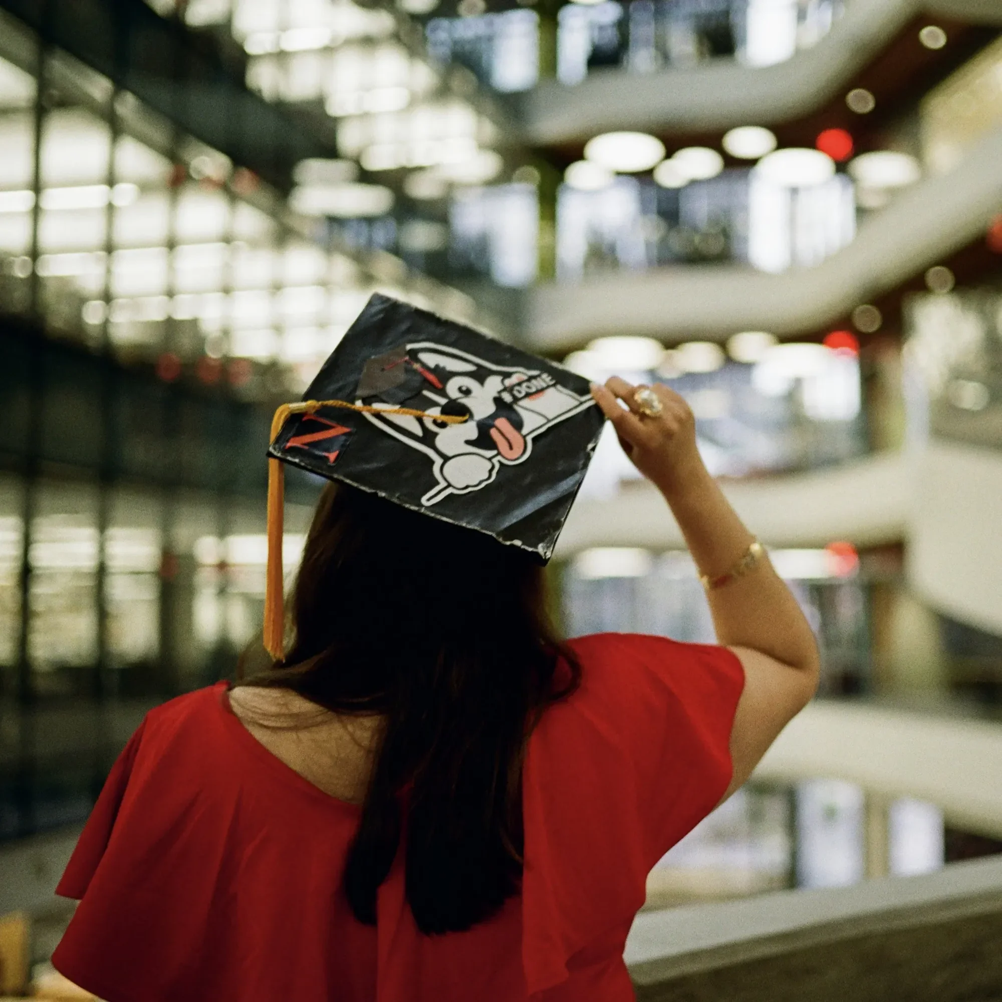 Person wearing a red dress holding graduation cap with a cartoon character design in a modern multi-story building.