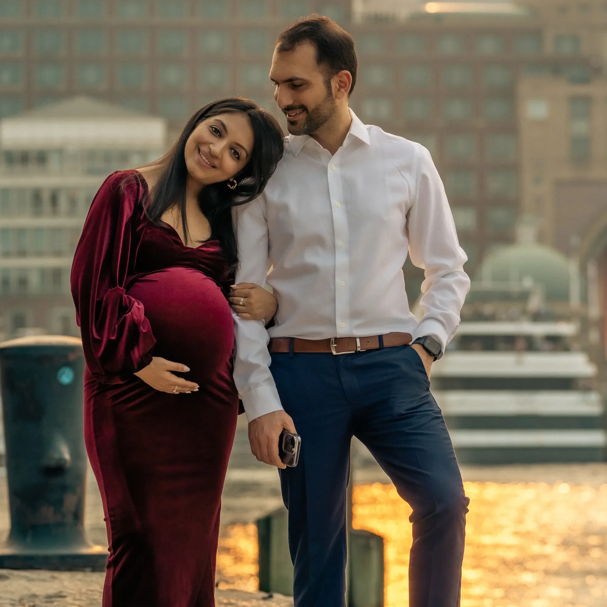 A pregnant woman in a burgundy velvet dress with long dark hair standing beside a man in white shirt and navy pants, both smiling and posing outdoors near a river with city buildings in the background at sunset.
