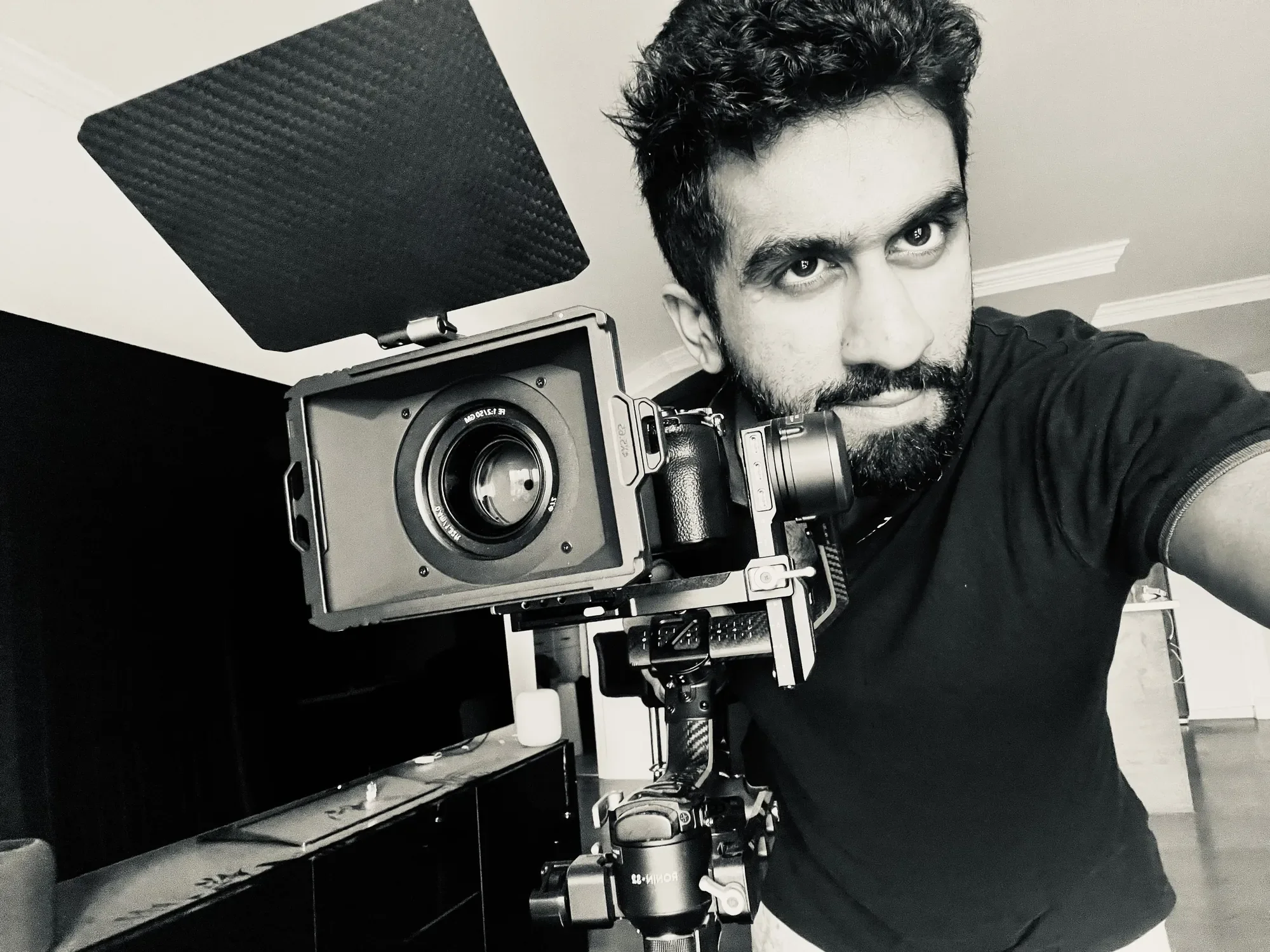 A man with dark curly hair and a beard taking a selfie, with a professional camera mounted on a stabilizer in front of him. The room has a ceiling with crown molding and a cabinet with a white object on top, with a predominantly black and white interior.