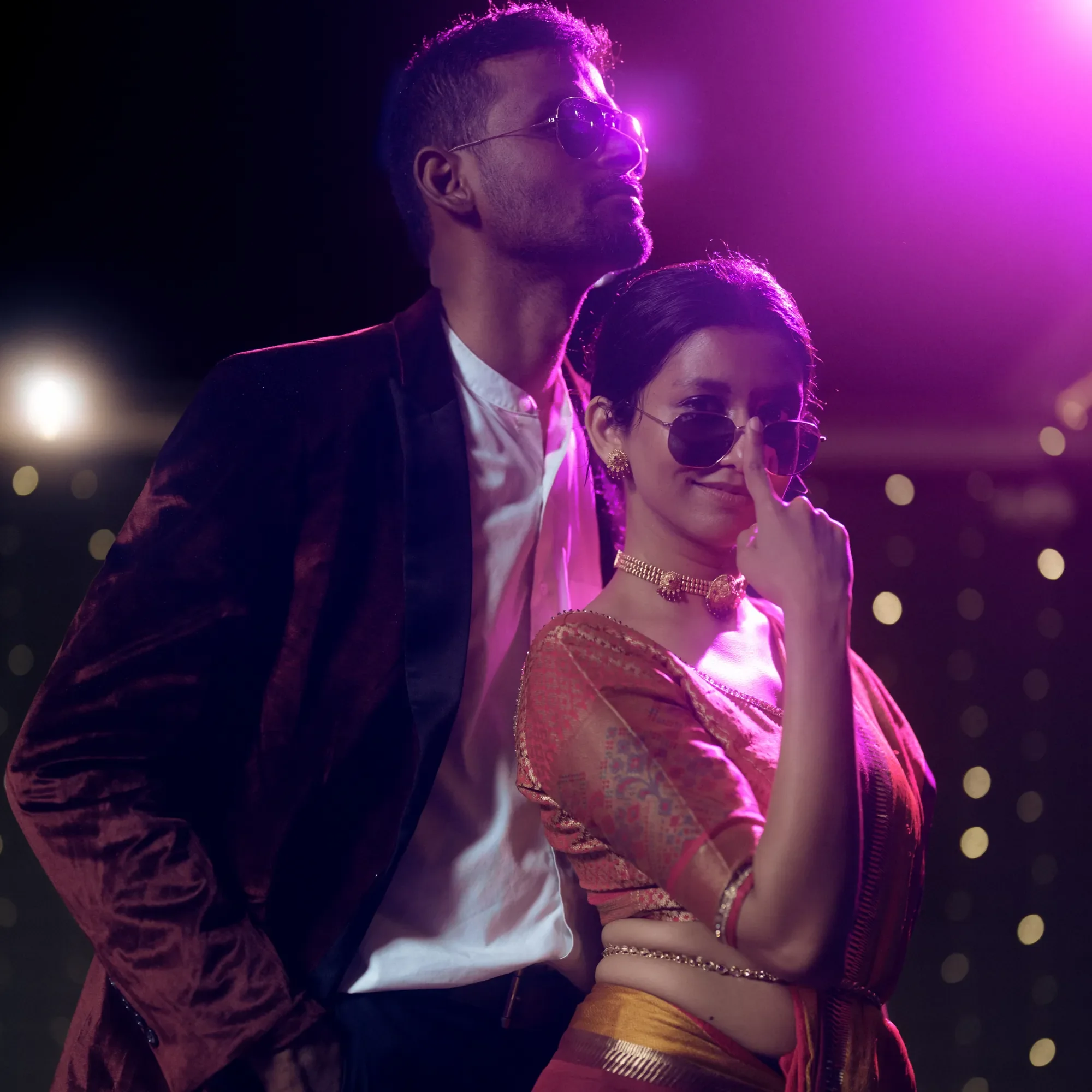 A man and woman wearing sunglasses and stylish clothing, standing together with a purple-lit background, with the woman making a finger gesture near her face.