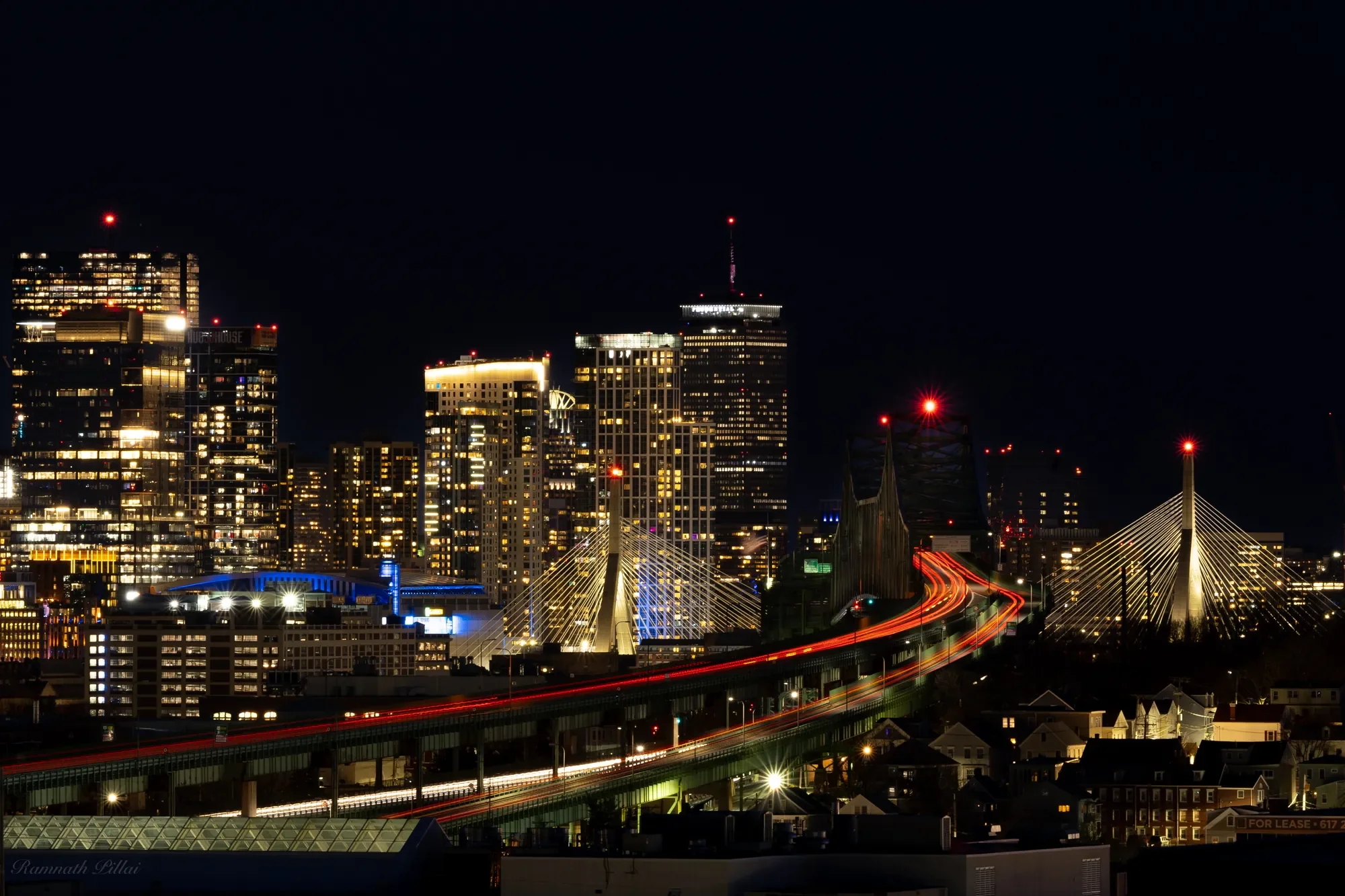Nighttime view of a city skyline with illuminated skyscrapers and bridges with light trails from moving vehicles.