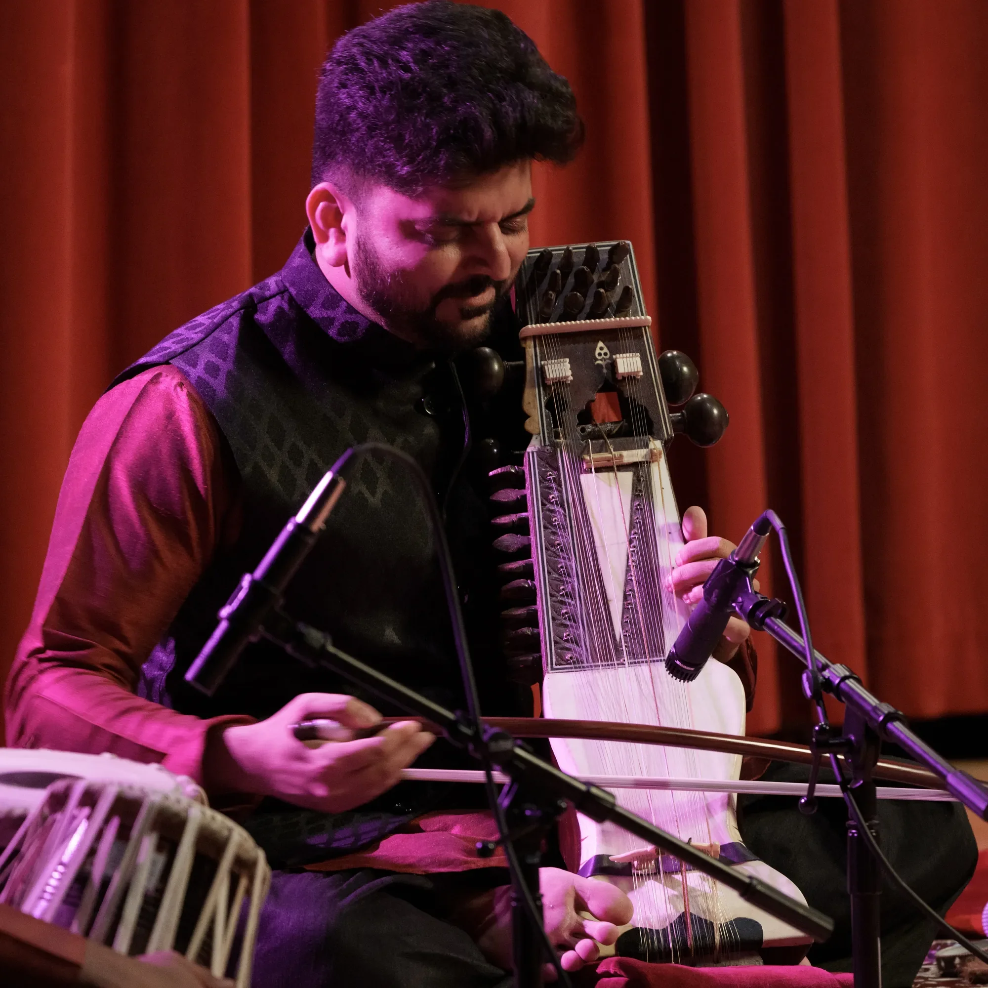 A man plays a traditional stringed instrument on stage, surrounded by microphones, with red curtains in the background.