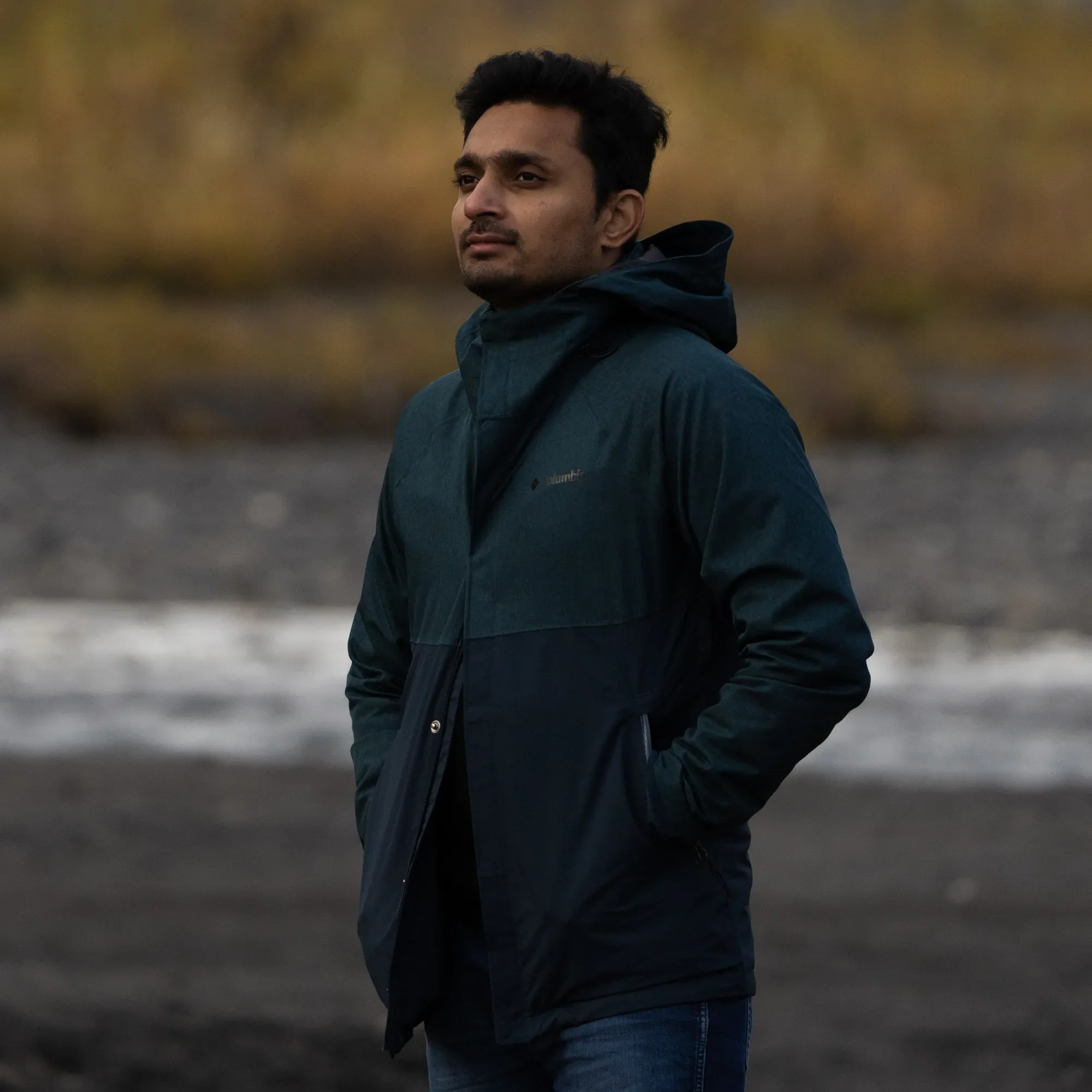 A man wearing a dark blue jacket with his hands in his pockets, standing outdoors near a body of water with blurred background of brownish foliage.