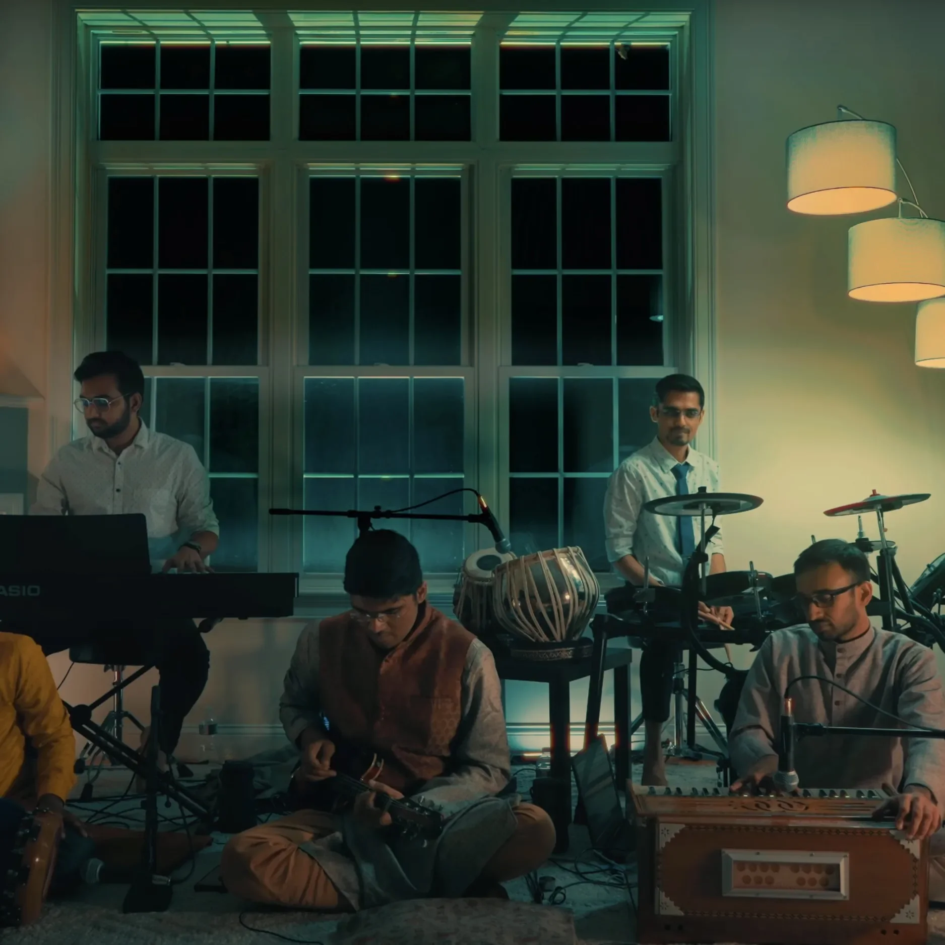 A band of five male musicians performing indoors at night, with large windows in the background and modern hanging lamps, playing various instruments including keyboard, guitar, drums, and harmonium.