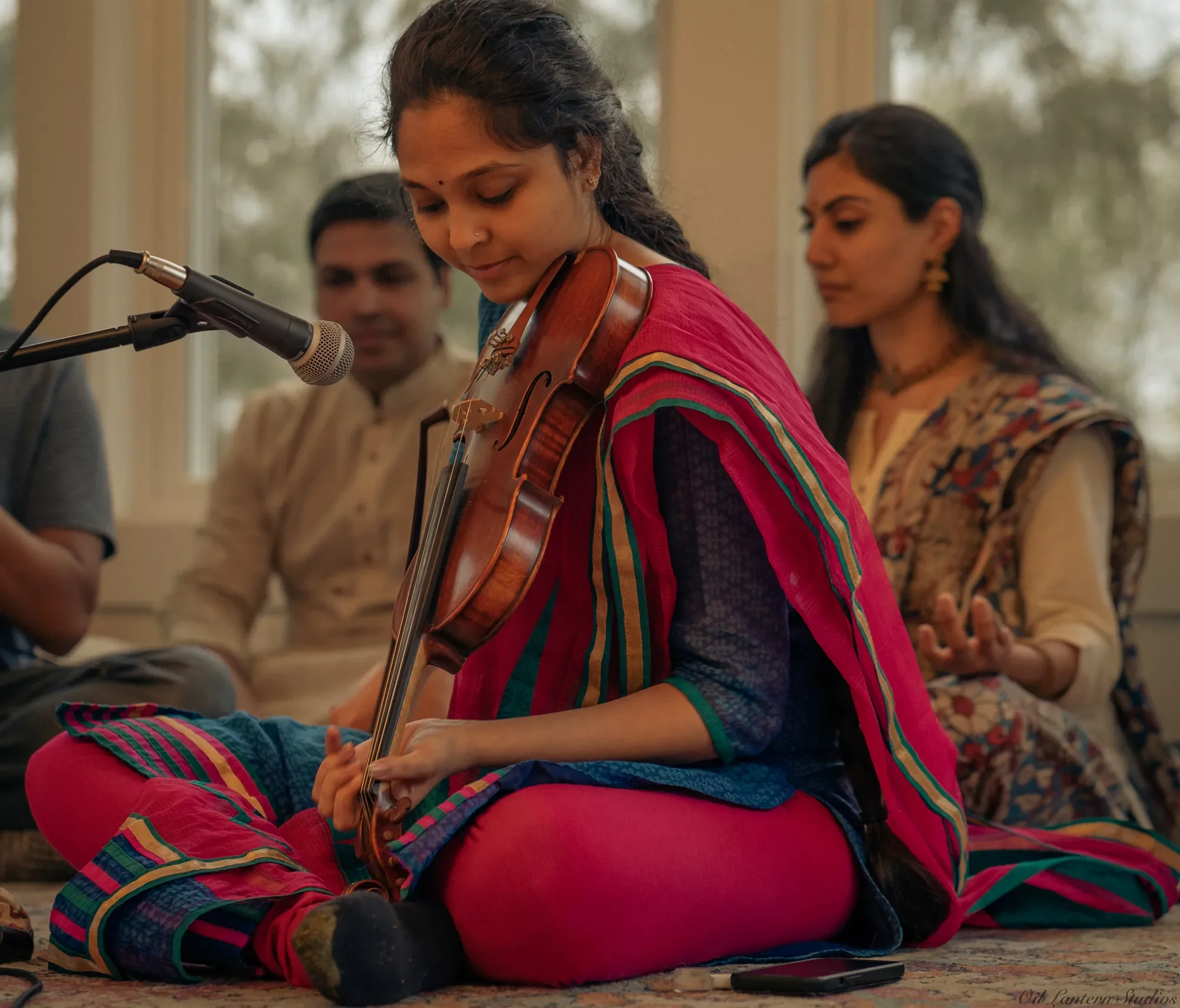 Young woman playing a violin during a traditional Indian music performance, sitting cross-legged on a carpet in front of a microphone, with two adults in traditional Indian attire in the background.
