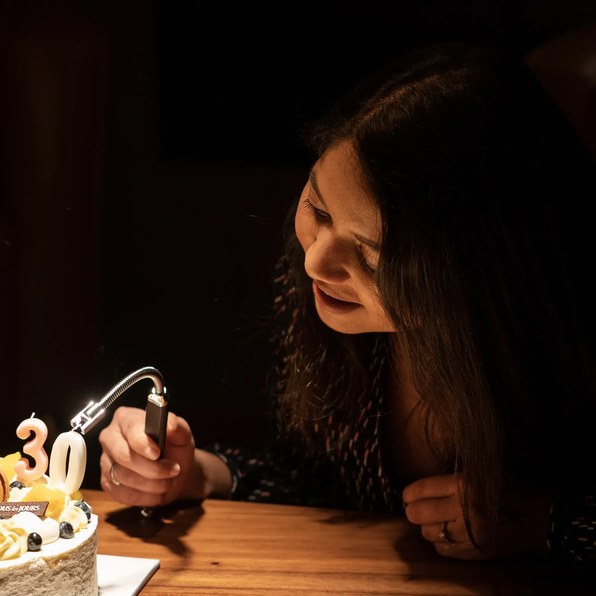 A woman with long dark hair is celebrating her 30th birthday, blowing out candles on a birthday cake with the number 30 shaped candles. The scene is dimly lit, and she is focused on the cake.