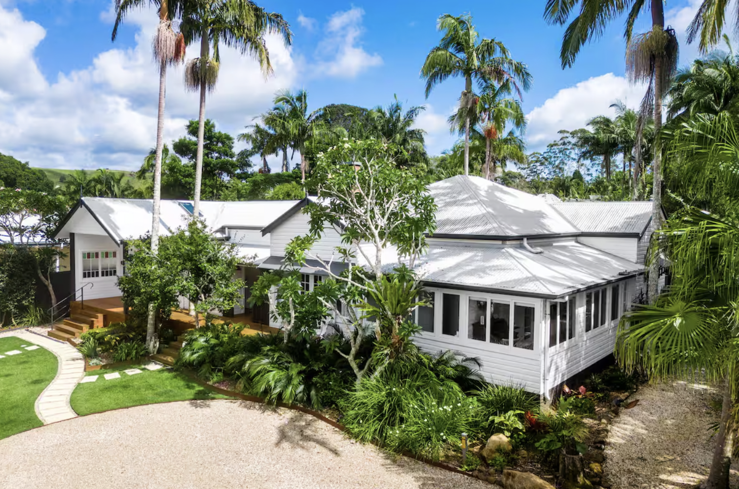 A white house with multiple roofs surrounded by lush greenery and tall palm trees, under a partly cloudy sky.
