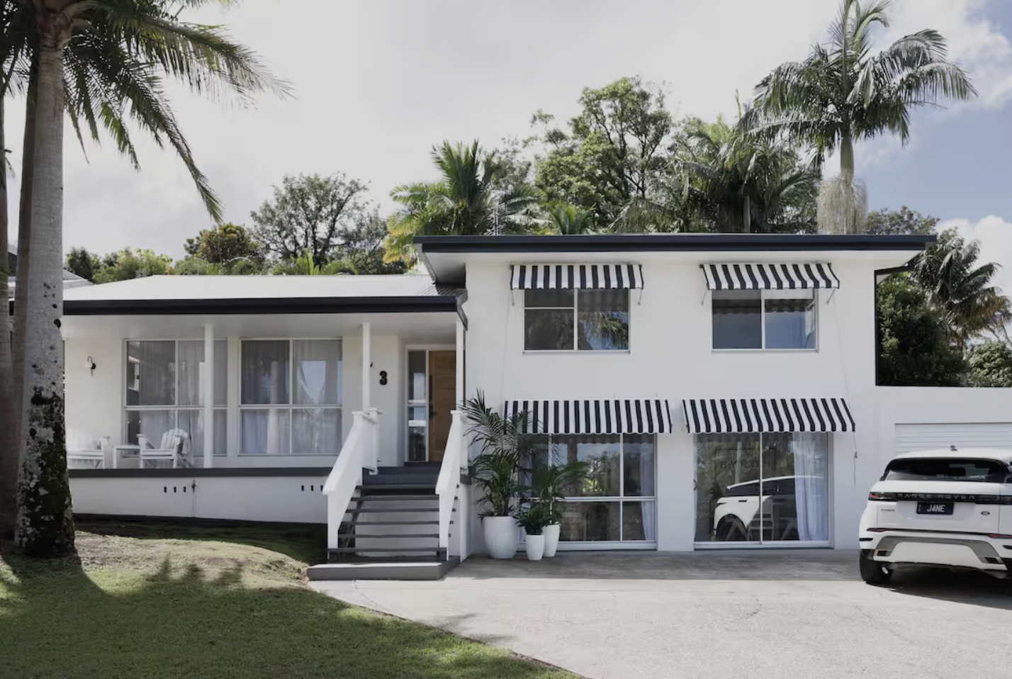 Two-story white house with black and white striped awnings over windows, front porch with white chairs, potted plants, driveway with white SUV, and surrounded by trees and greenery.