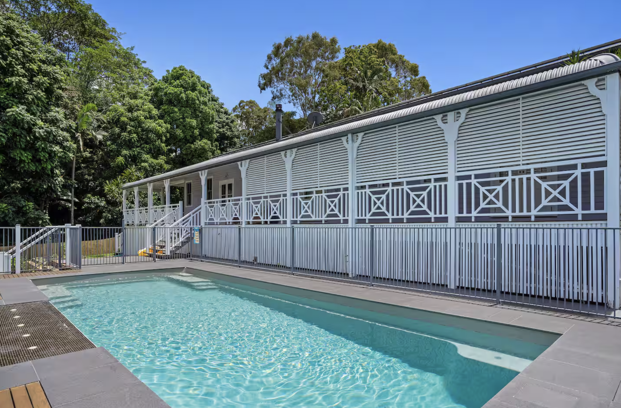 A backyard with a swimming pool, a gray house with a white porch, surrounded by green trees under a clear blue sky.