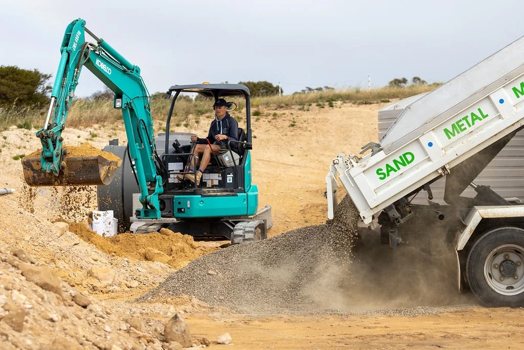 A person operating a small blue excavator on a construction site, moving dirt or sand, with a truck dumping a mixture of sand and gravel labeled 'SAND', 'METAL', and 'M...'
