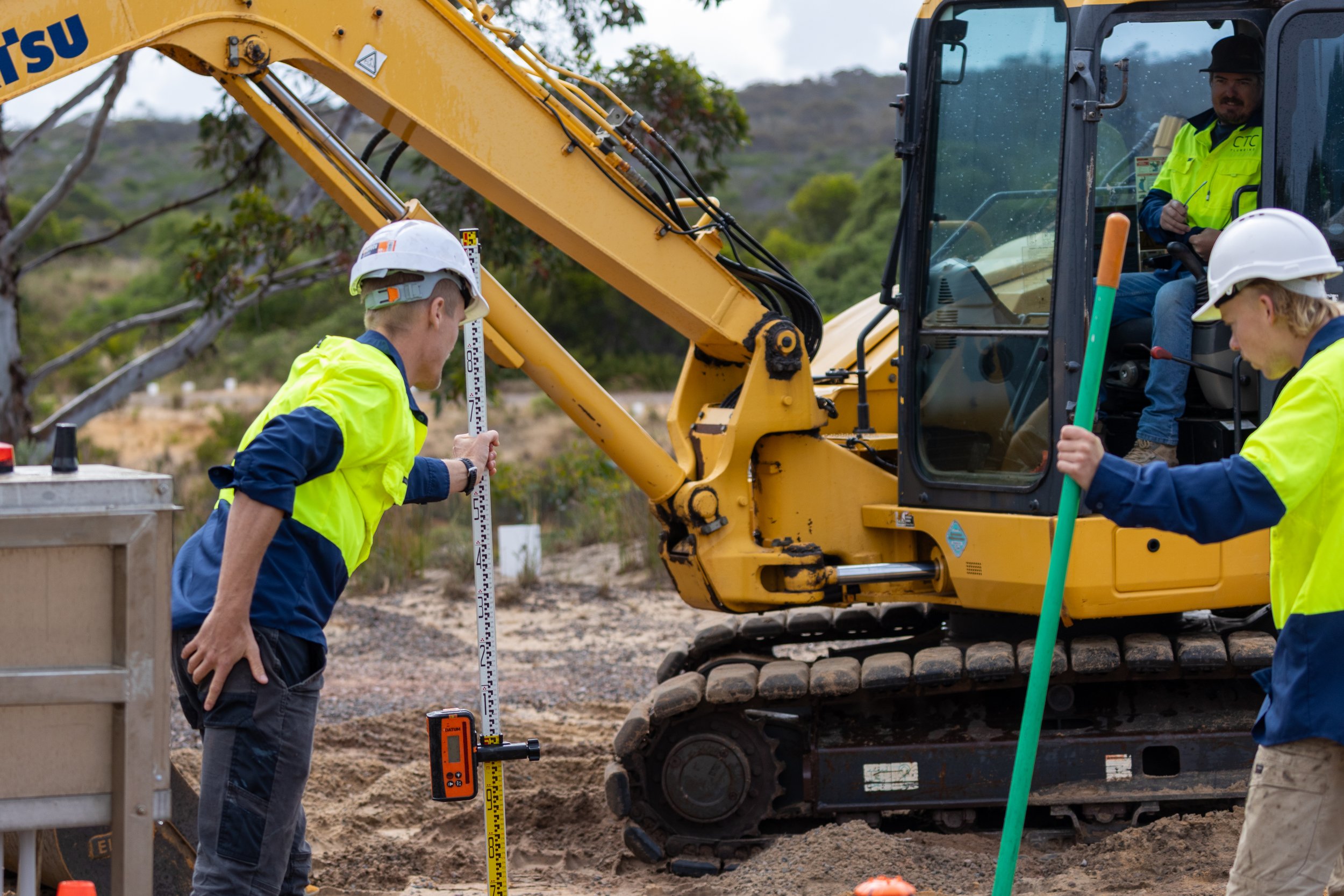 Person at a construction site holding a measuring rod, with surveying equipment and pipes nearby under a partly cloudy sky.