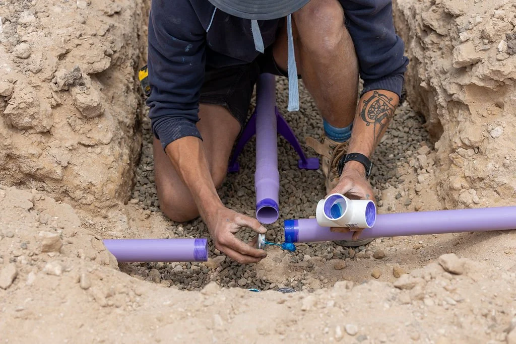 A person working on installing purple PVC pipes in a dug trench outside.