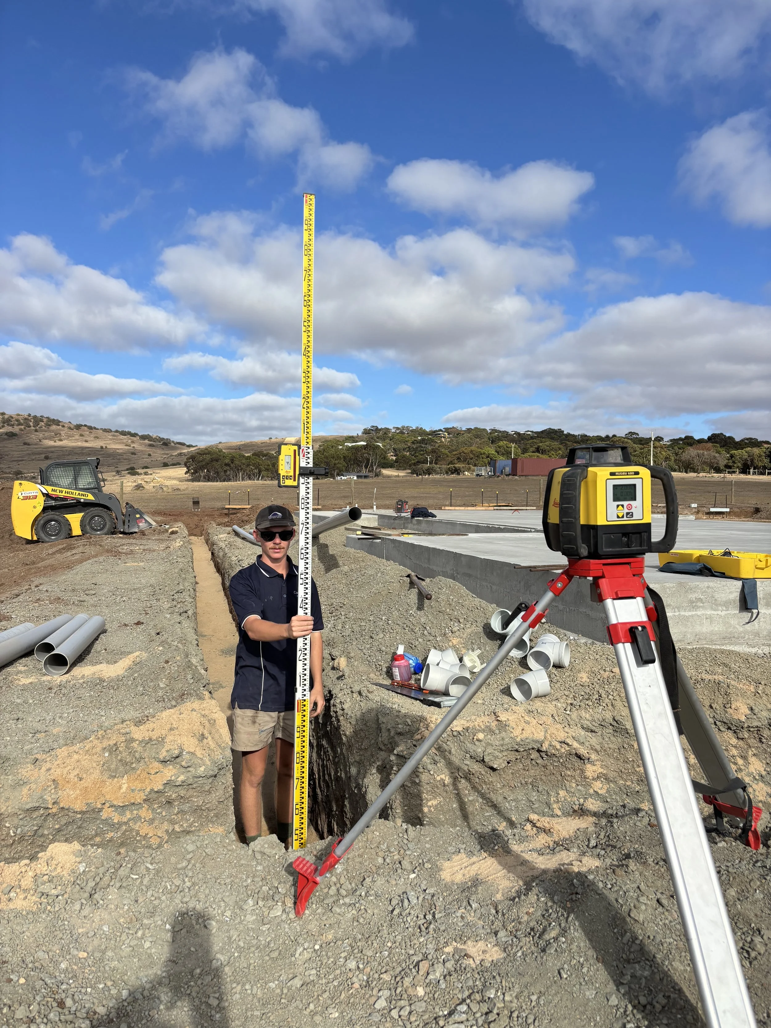 Person at a construction site holding a measuring rod, with surveying equipment and pipes nearby under a partly cloudy sky.