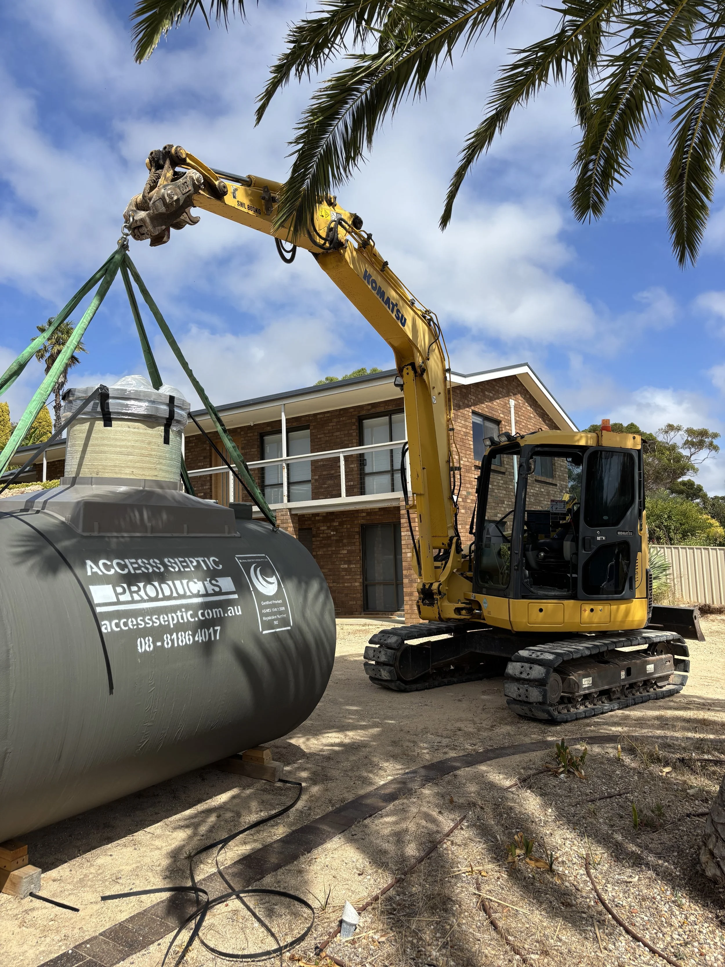 A small yellow excavator lifting a large black septic tank onto the ground at a construction site in front of a residential building with a balcony and brick exterior.
