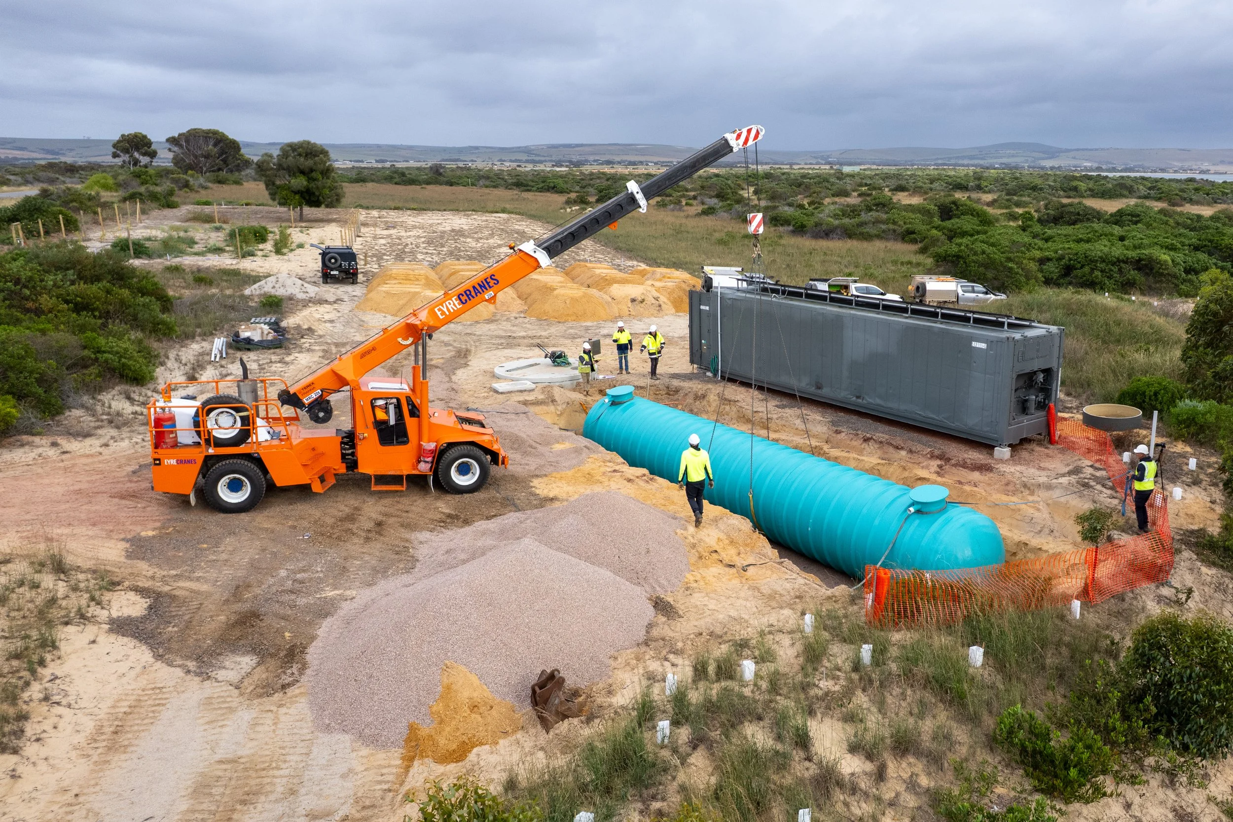 A person operating a small blue excavator on a construction site, moving dirt or sand, with a truck dumping a mixture of sand and gravel labeled 'SAND', 'METAL', and 'M...'