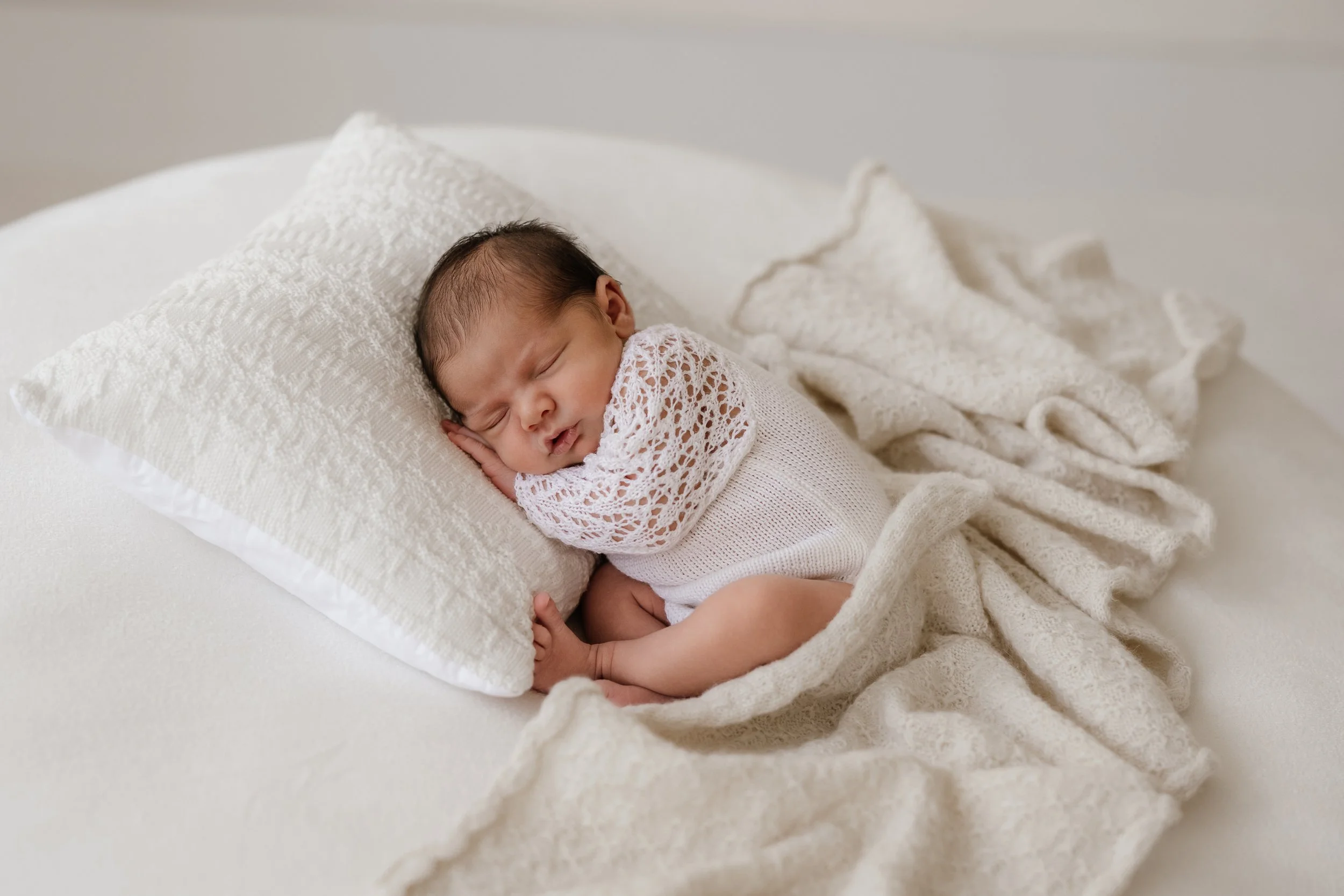 A newborn baby sleeping on a cream-colored bed with a textured pillow and cozy blanket, dressed in a white crochet top.