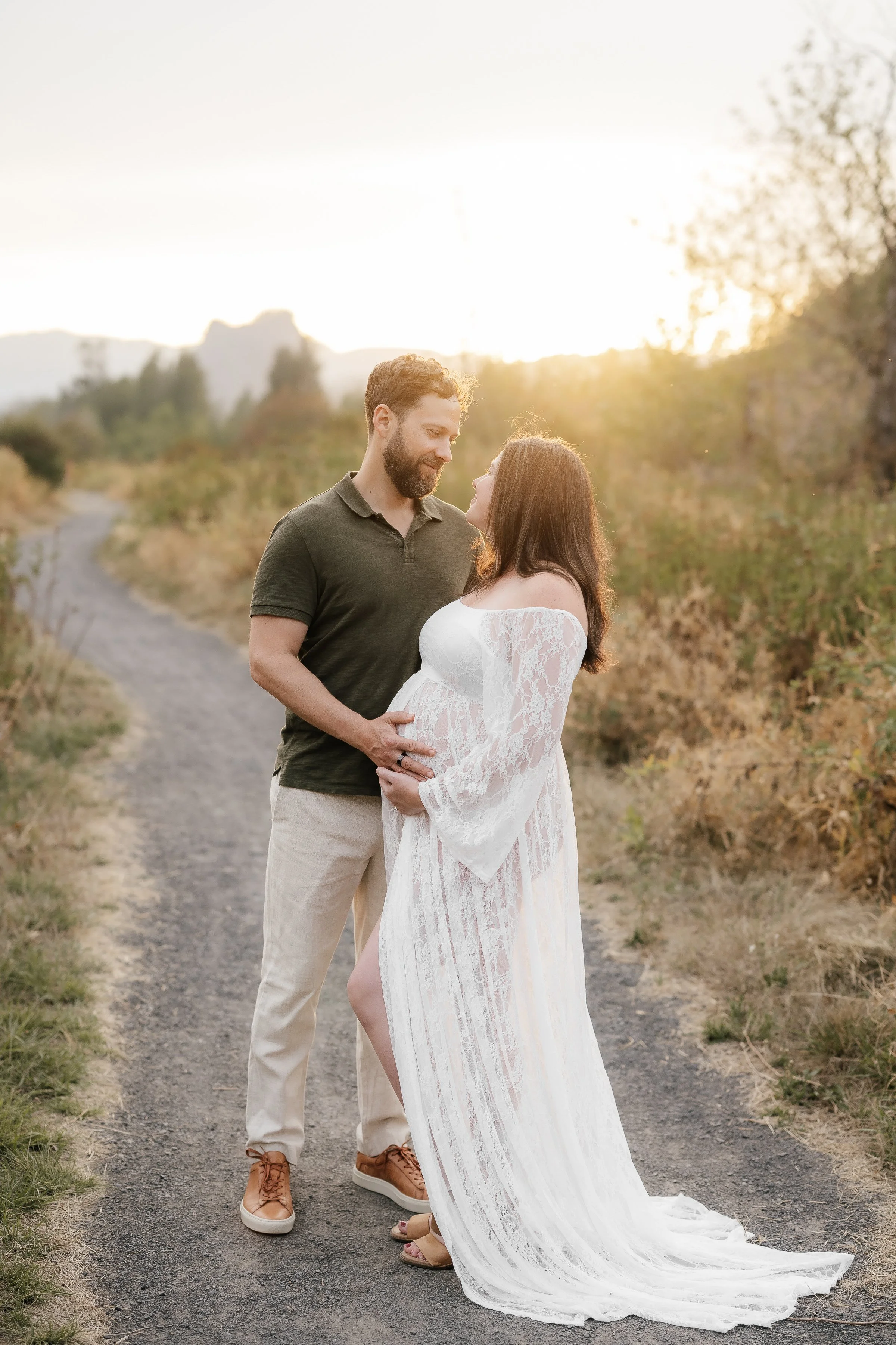 A pregnant woman and a man sharing an intimate moment outdoors on a dirt path during sunset. The woman is wearing a long, white lace dress, and the man is dressed in a dark polo shirt and light-colored pants.