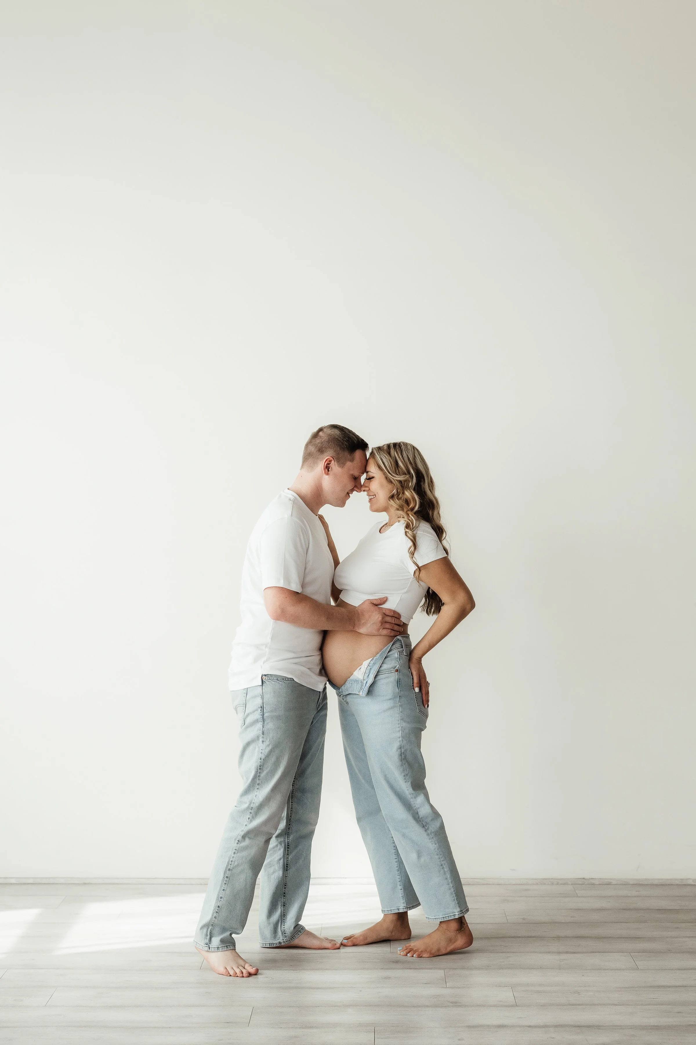 A pregnant woman and a man, both in casual white shirts and jeans, standing barefoot on a light-colored wooden floor, with foreheads touching and holding her belly affectionately against a plain white wall.