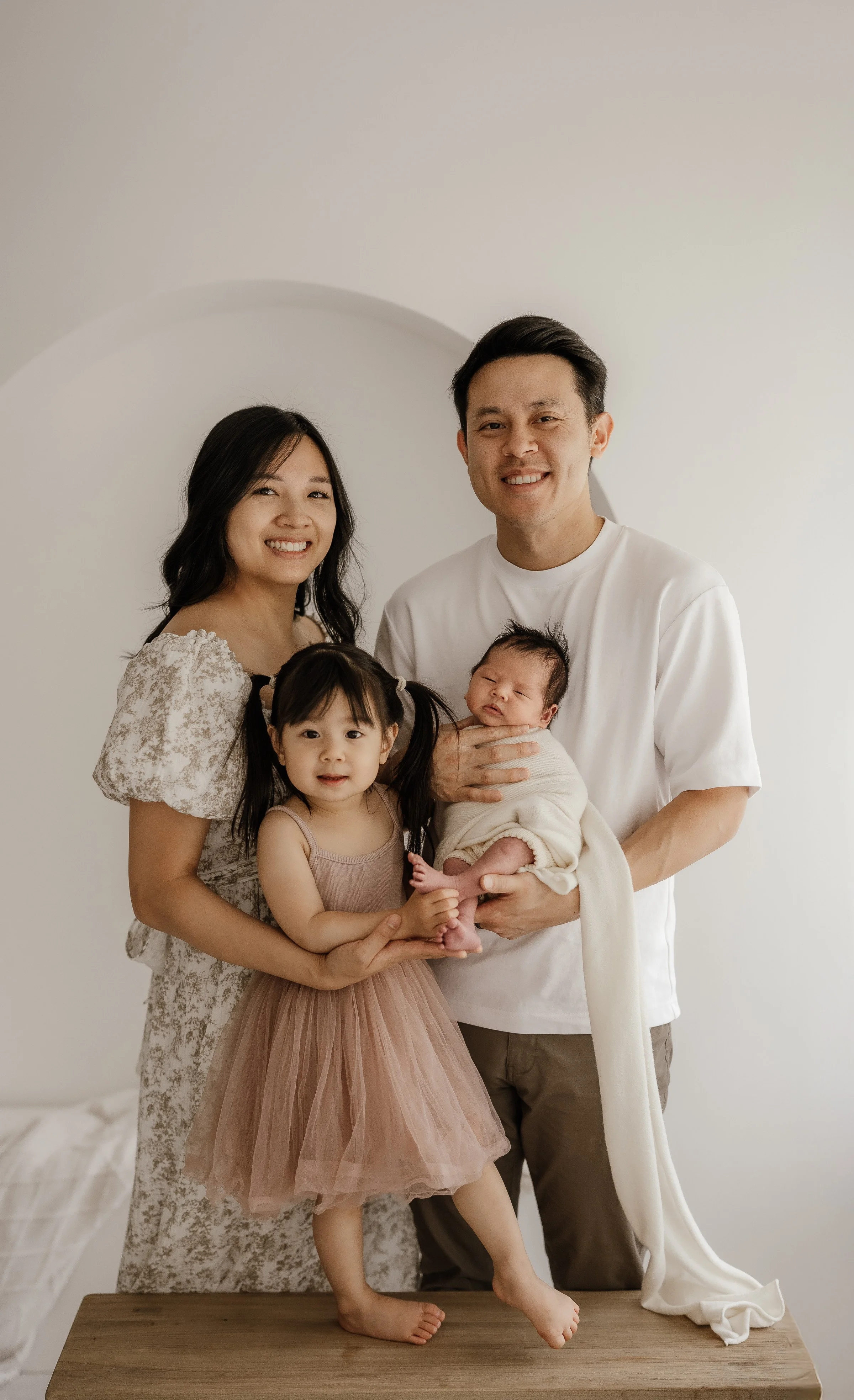 Family portrait of a man, woman, and two young girls standing together in a neutral-colored room, with the man holding a newborn baby wrapped in a blanket.