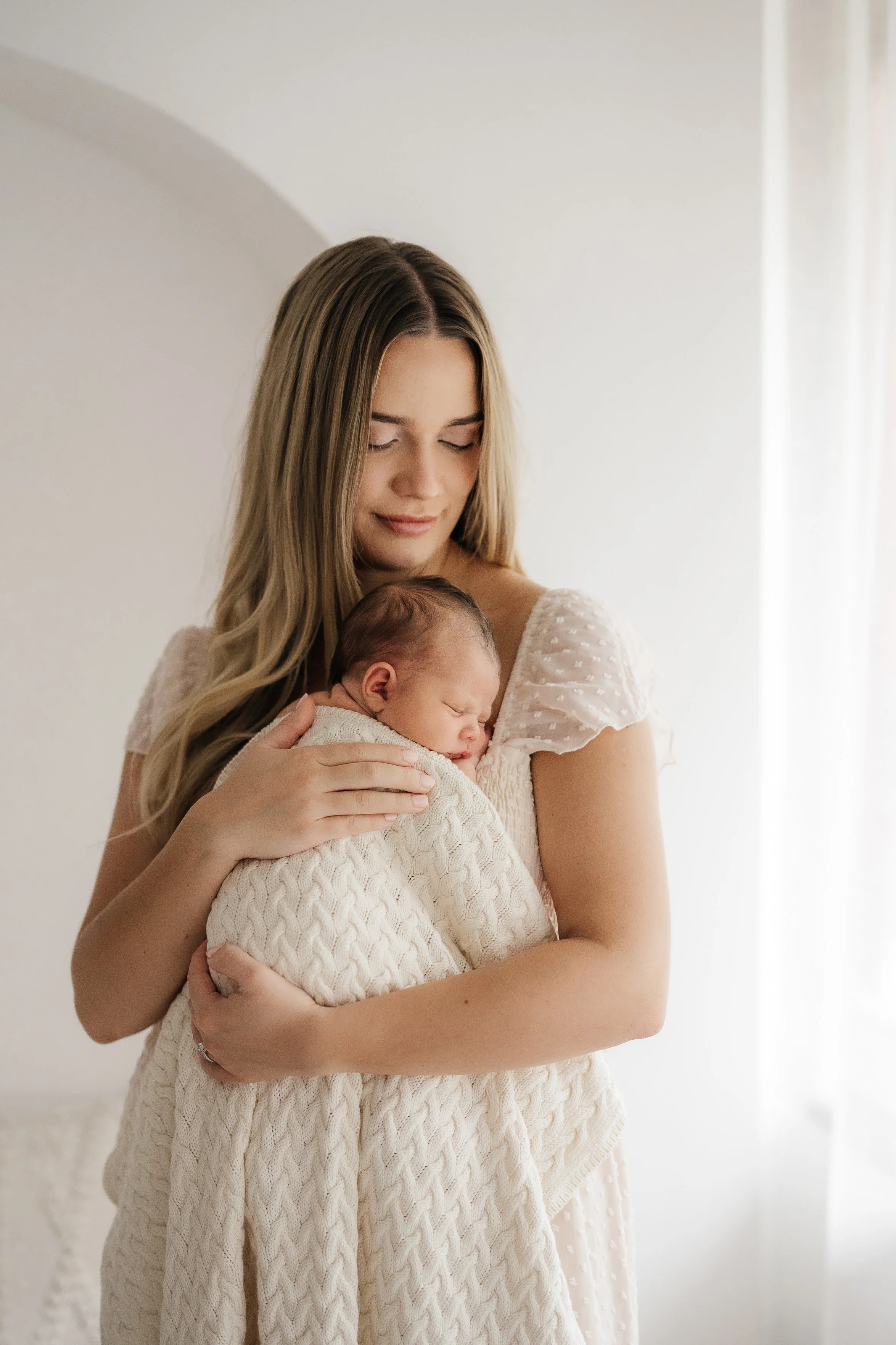 A young woman holding a sleeping baby wrapped in a knitted blanket in a softly lit room.