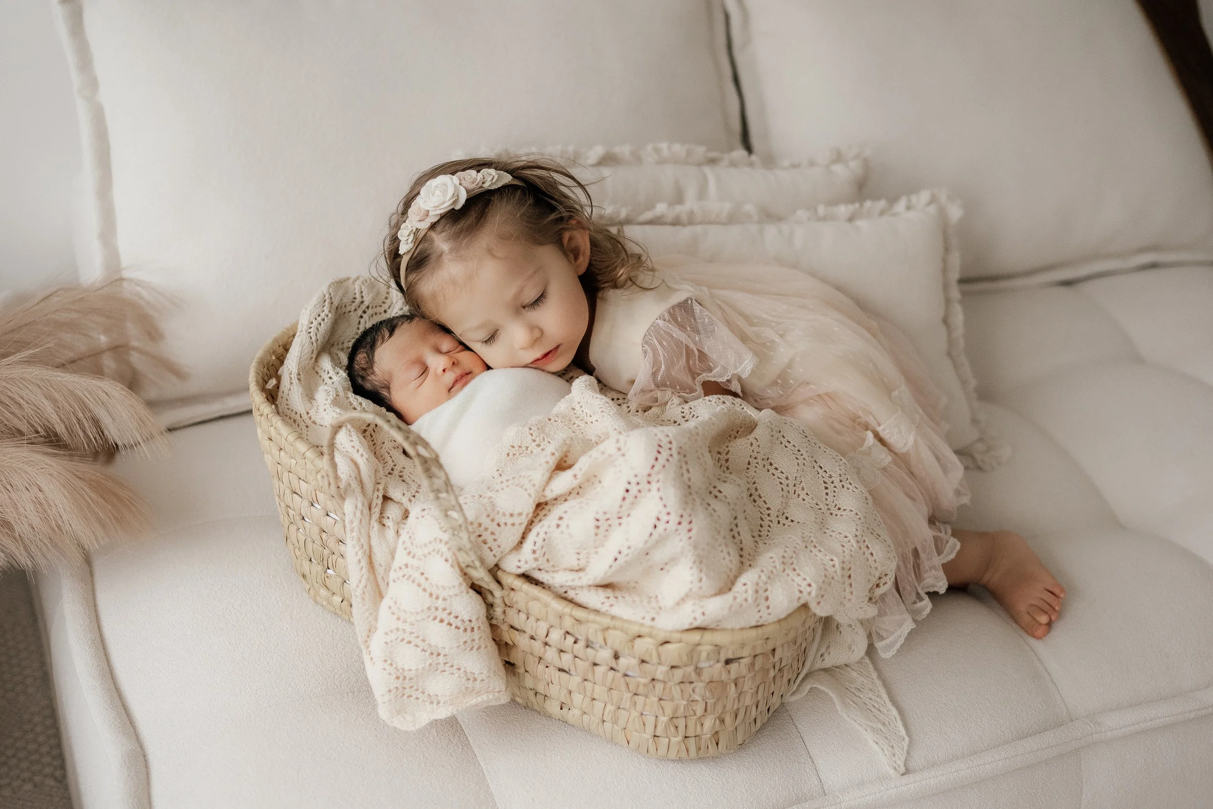 A young girl with a flower headband cuddling a newborn baby in a basket on a white sofa.