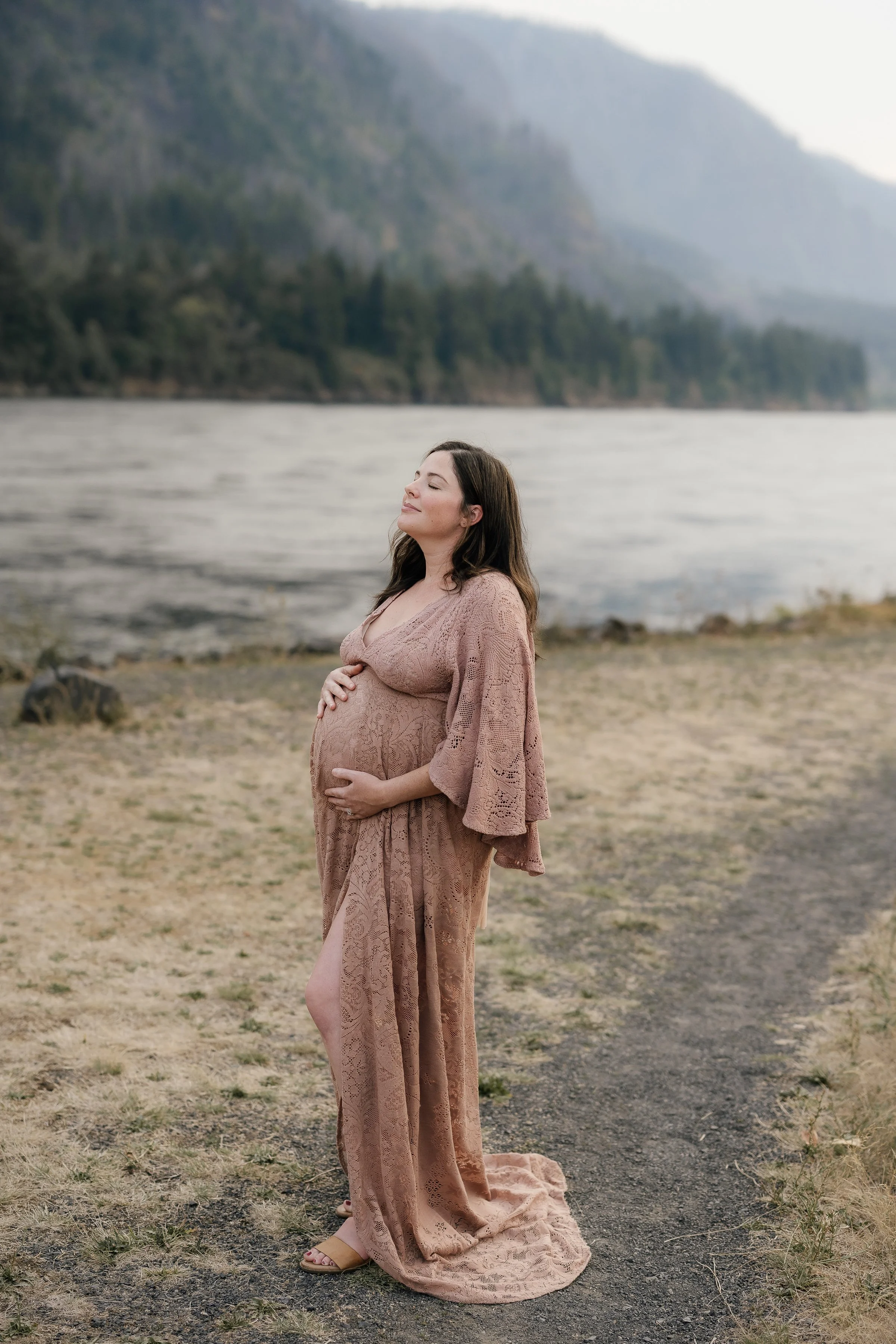 Pregnant woman in a pink lace dress standing on a natural landscape with water and mountains in the background, eyes closed, enjoying the moment.