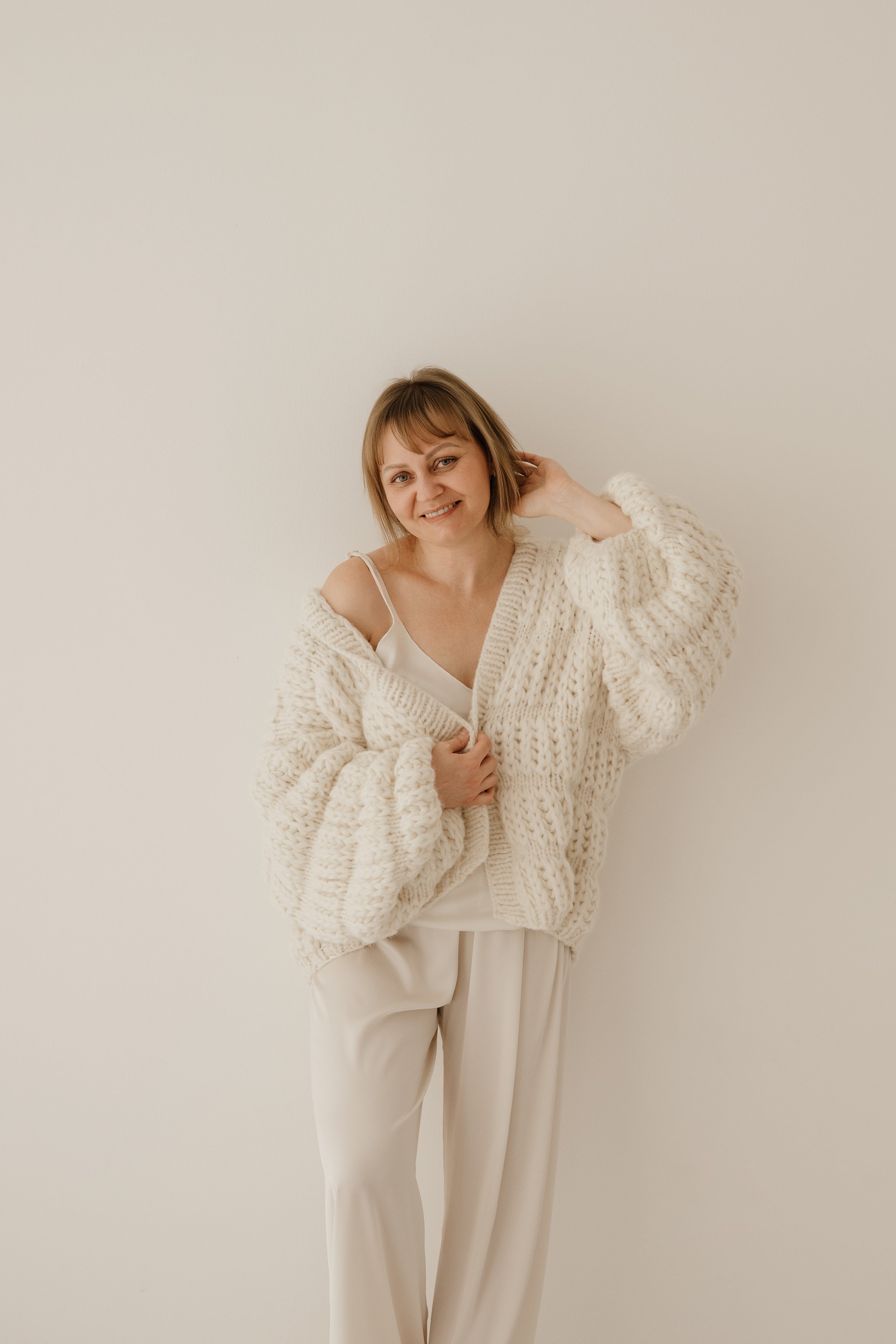 A woman standing against a the plain white wall wearing a cream-colored chunky knit sweater, a white camisole, and cream-colored trousers, smiling with her hand touching her head.