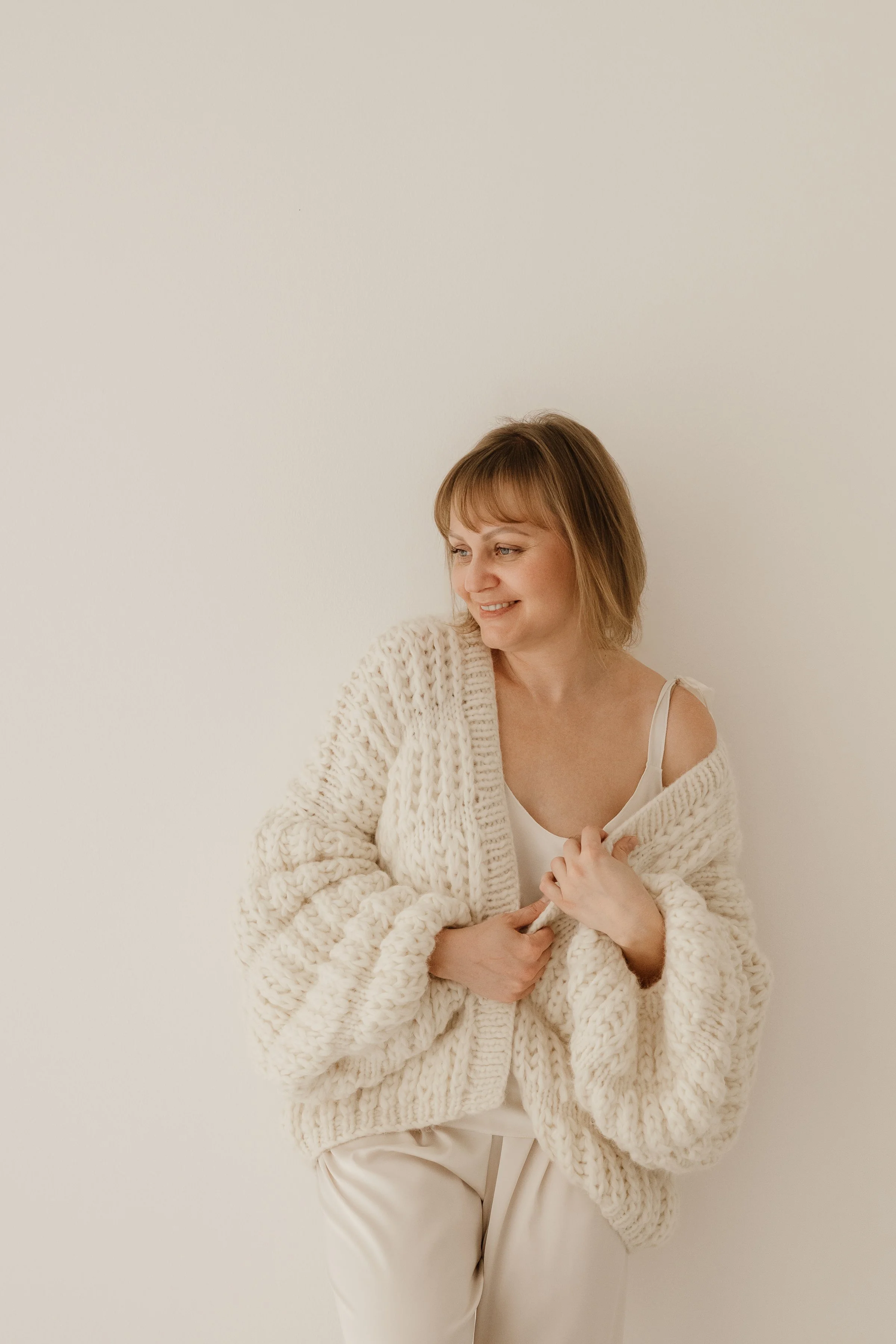 A woman with short light brown hair wearing a chunky cream knitted sweater over a white satin camisole, standing against a plain off-white background, smiling gently and looking to her left.