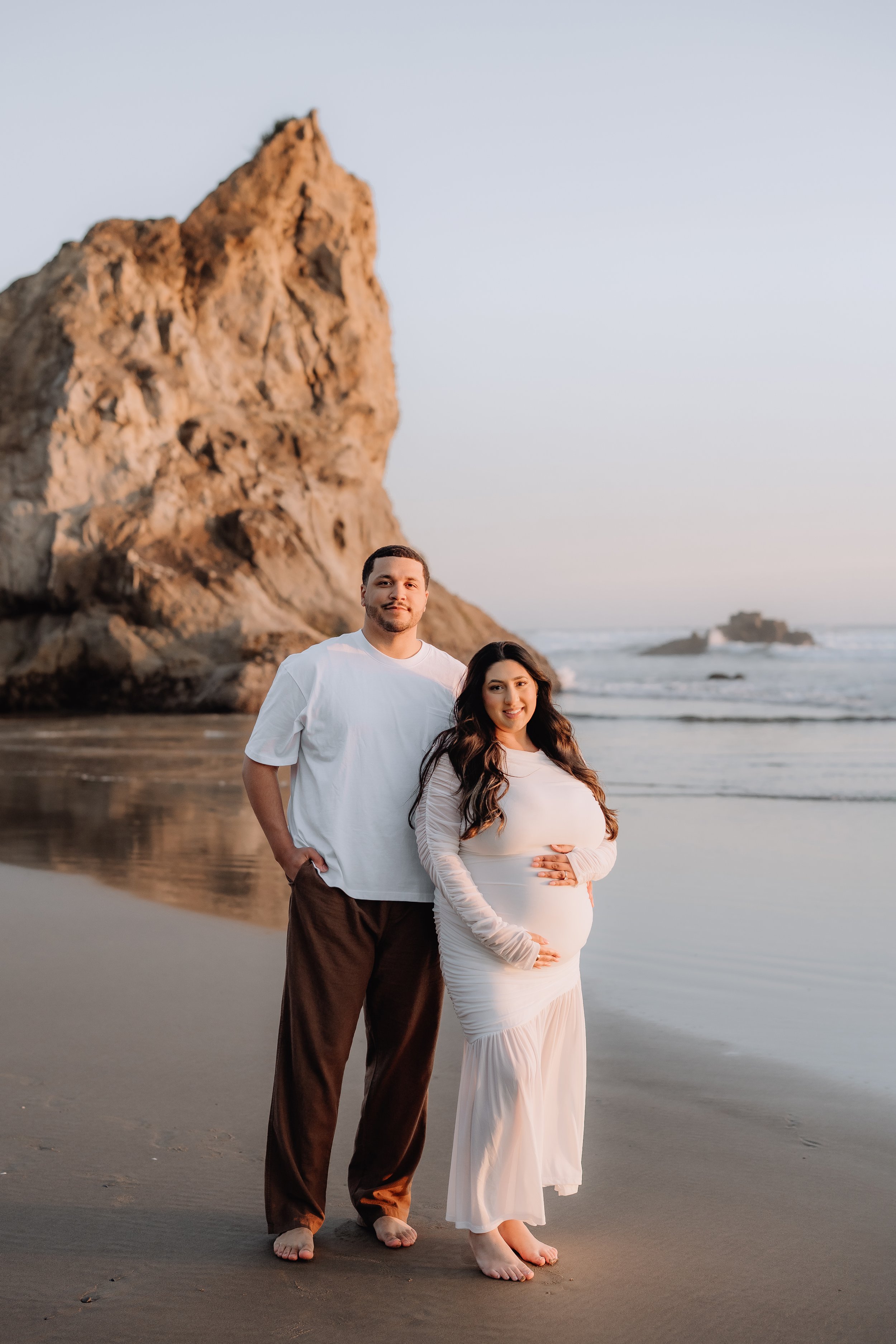 A pregnant woman and a man standing on a beach at sunset, with large rock formations and the ocean in the background.