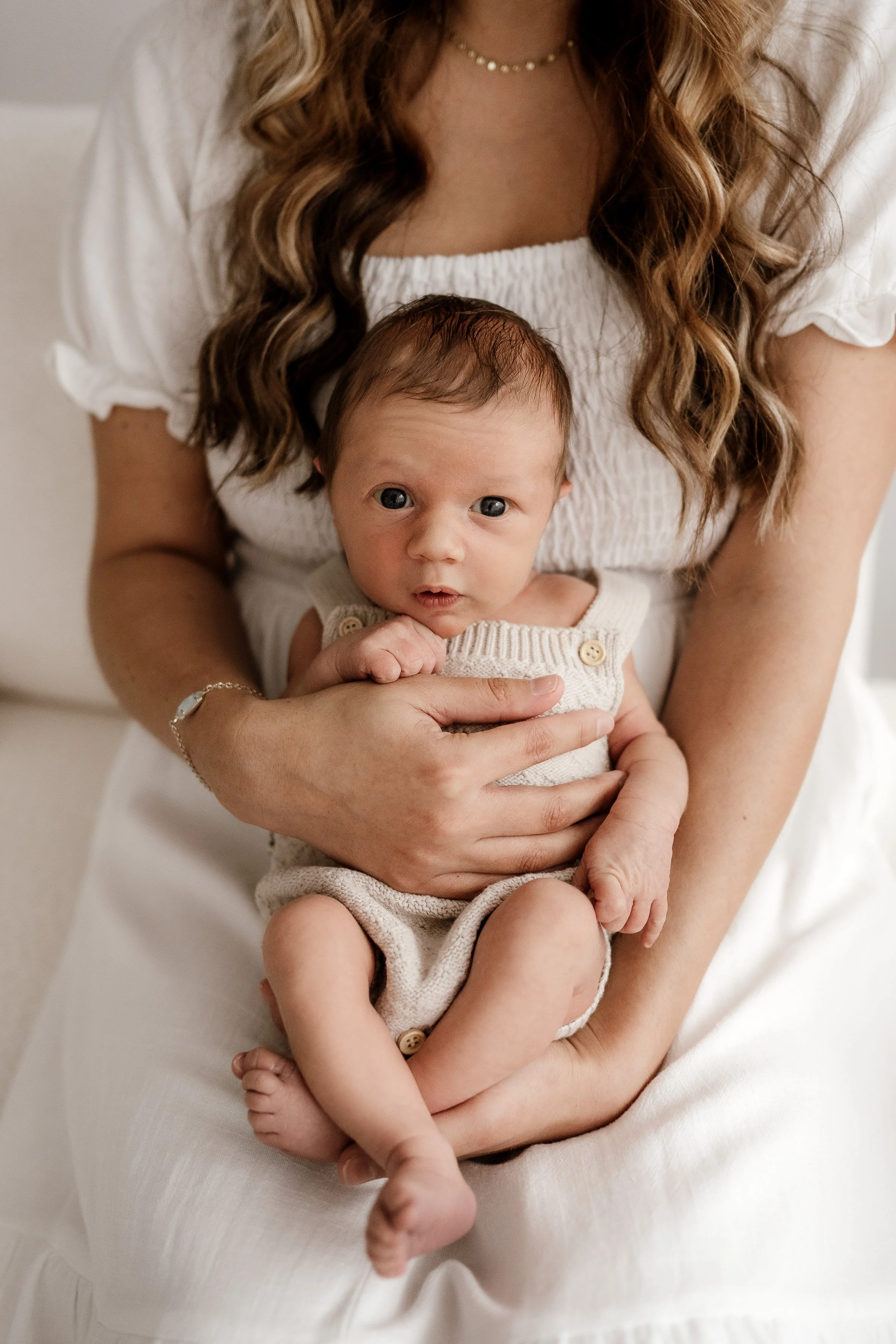 A woman with wavy brown hair wearing a white dress and a pearl necklace is holding a small baby with blue eyes and light brown hair, dressed in a beige knit outfit.