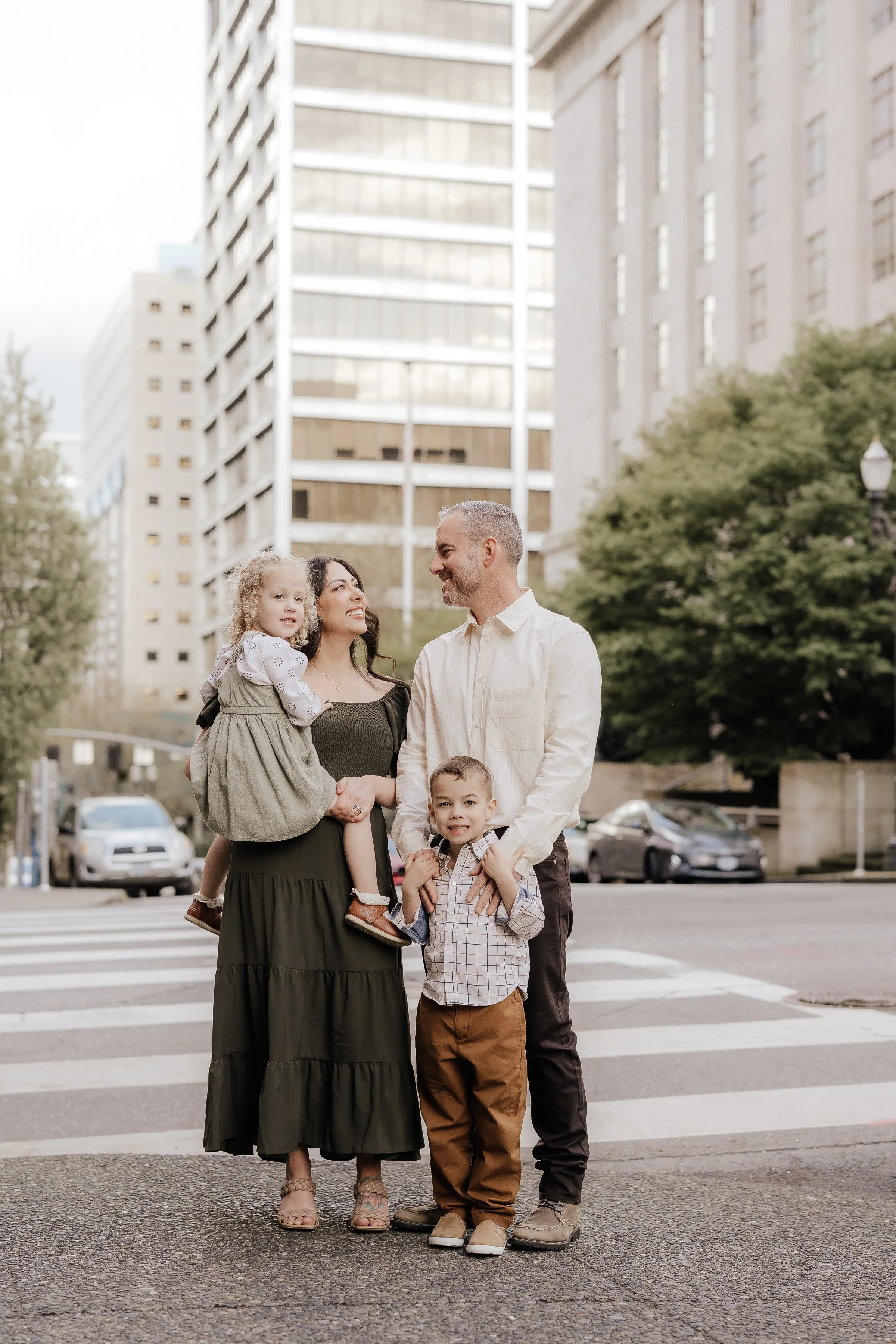 A family of four standing at a crosswalk in an urban area, with tall buildings and trees in the background.