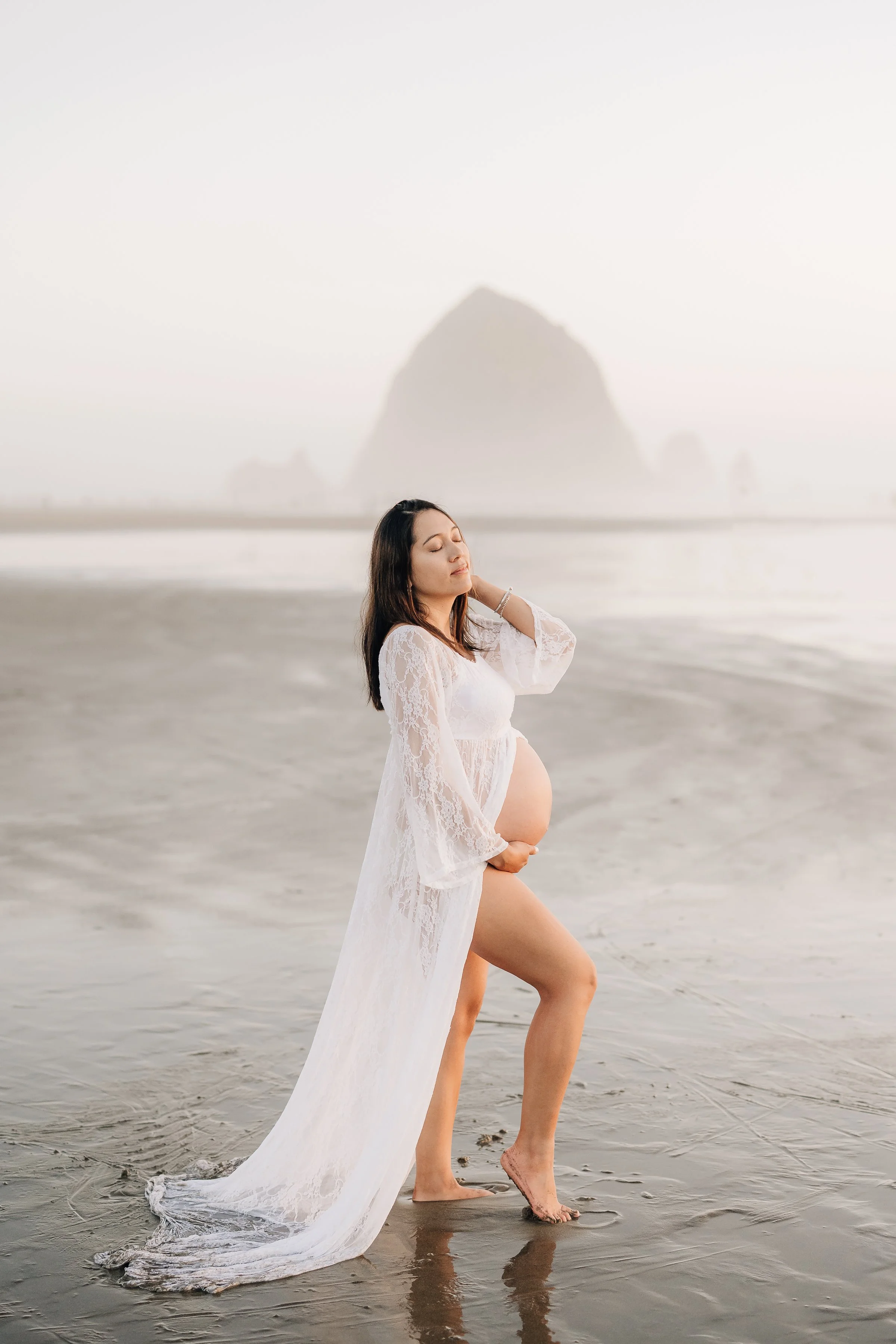 A pregnant woman in a white lace dress standing on a wet beach, with her eyes closed and one hand behind her head, and a large mountain in the background.