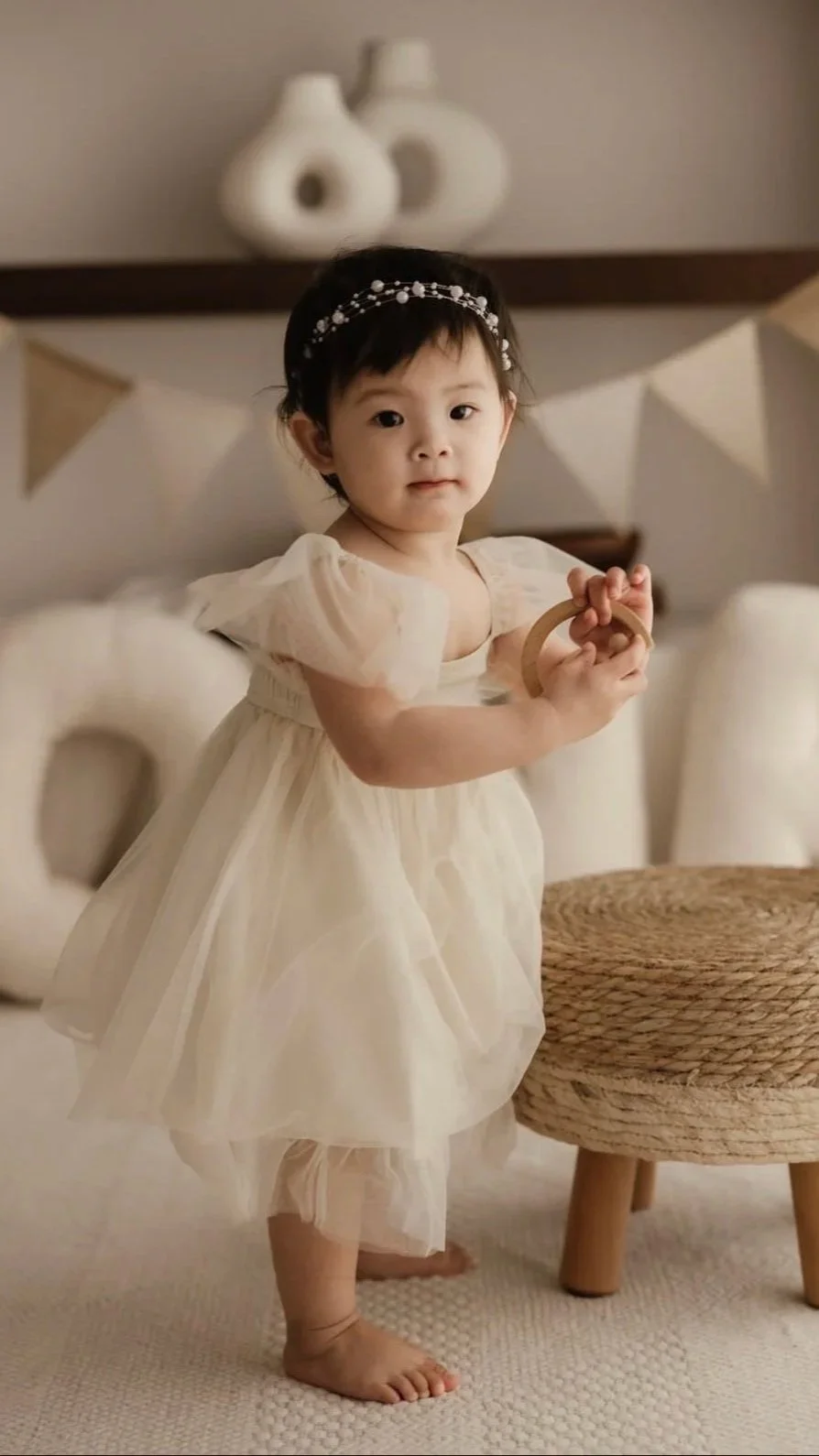A young girl in a cream-colored dress with puffed sleeves and a tulle skirt, barefoot, standing indoors with a neutral expression, holding a wooden ring. She has a delicate headband with small beads or pearls. In the background, there are decorative 