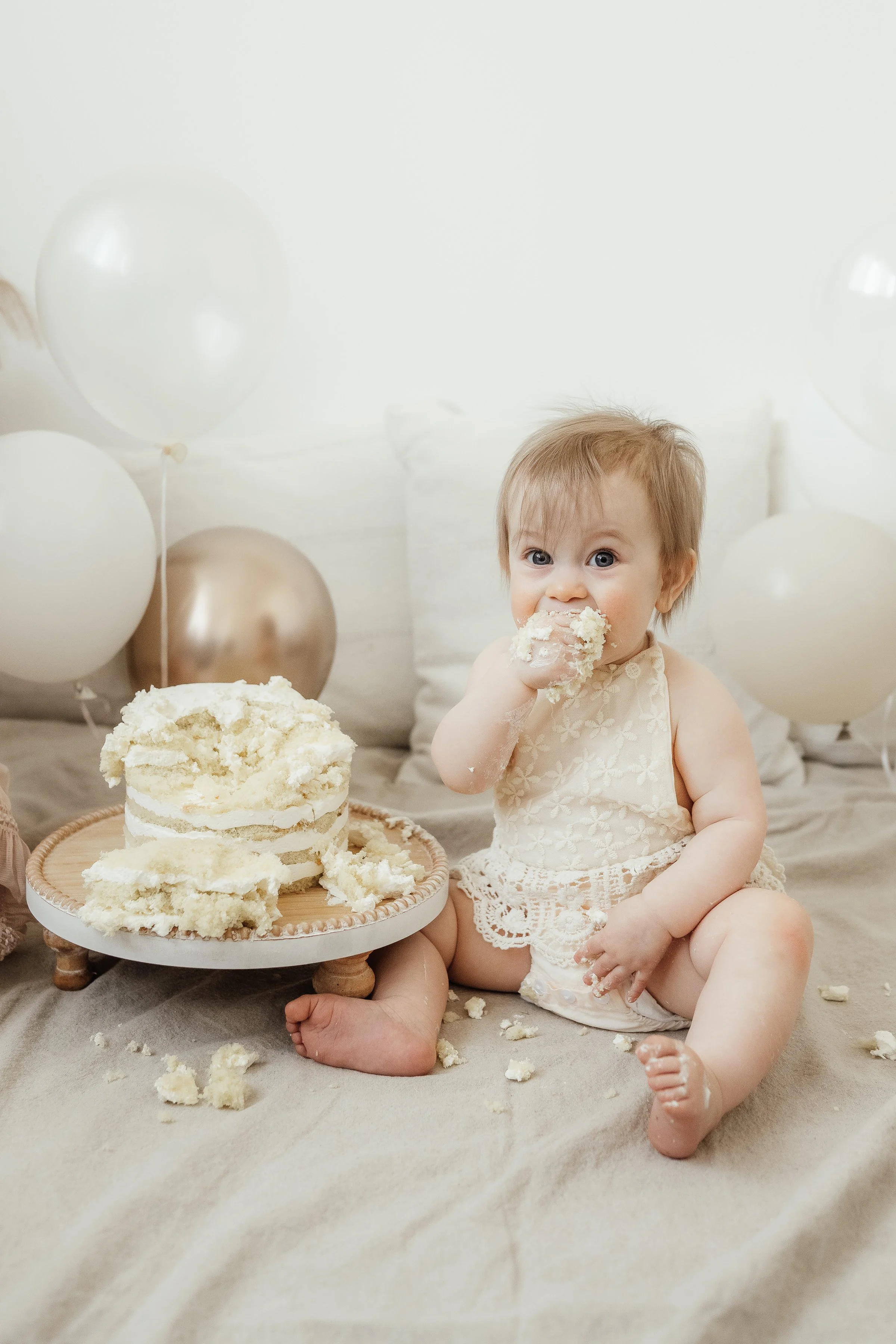 A baby girl in a cream dress playing with cake and frosting on a cake table, surrounded by white and gold balloons, in a celebration setting.