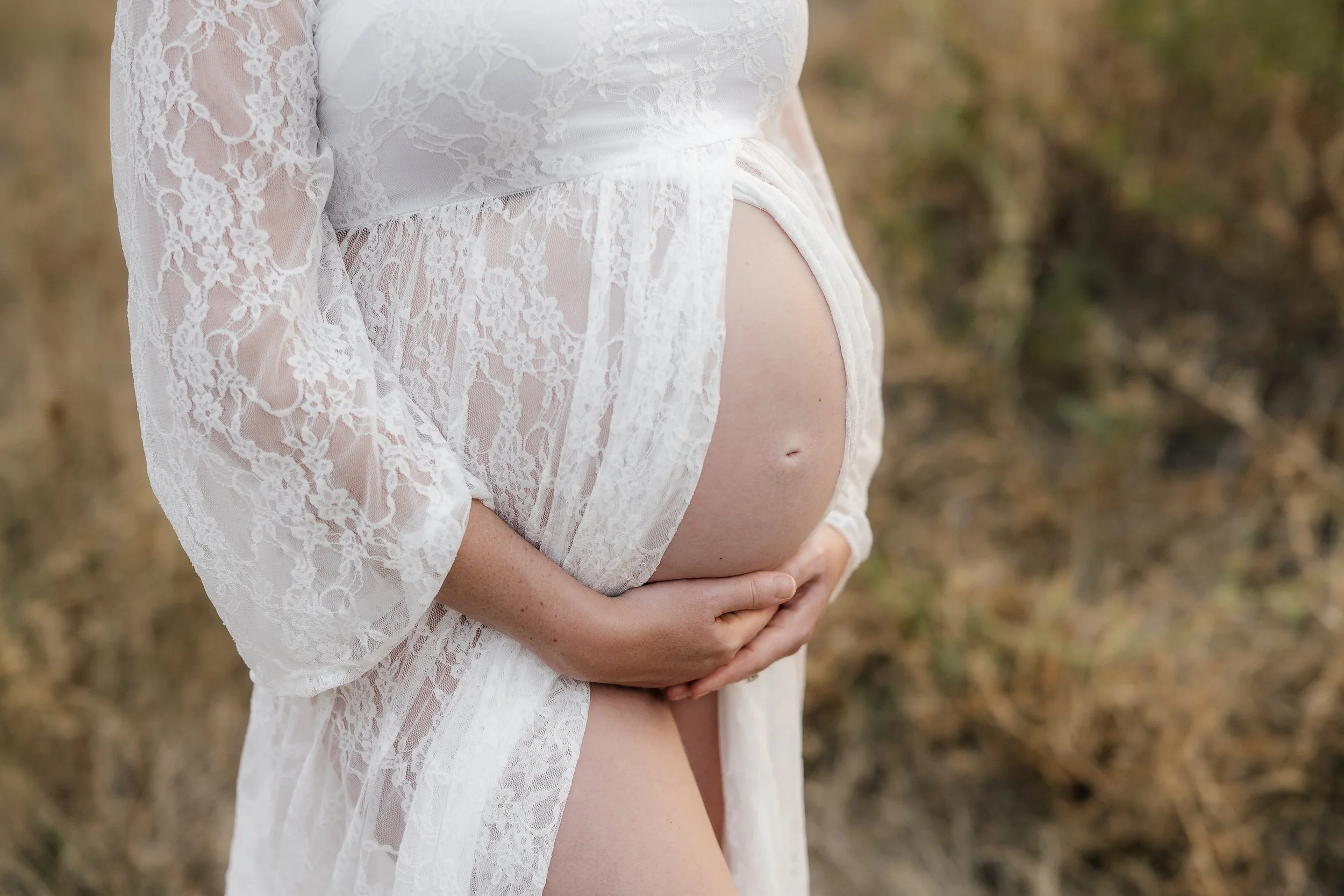 Close-up of a pregnant woman wearing a white lace dress, gently holding her belly outdoors.