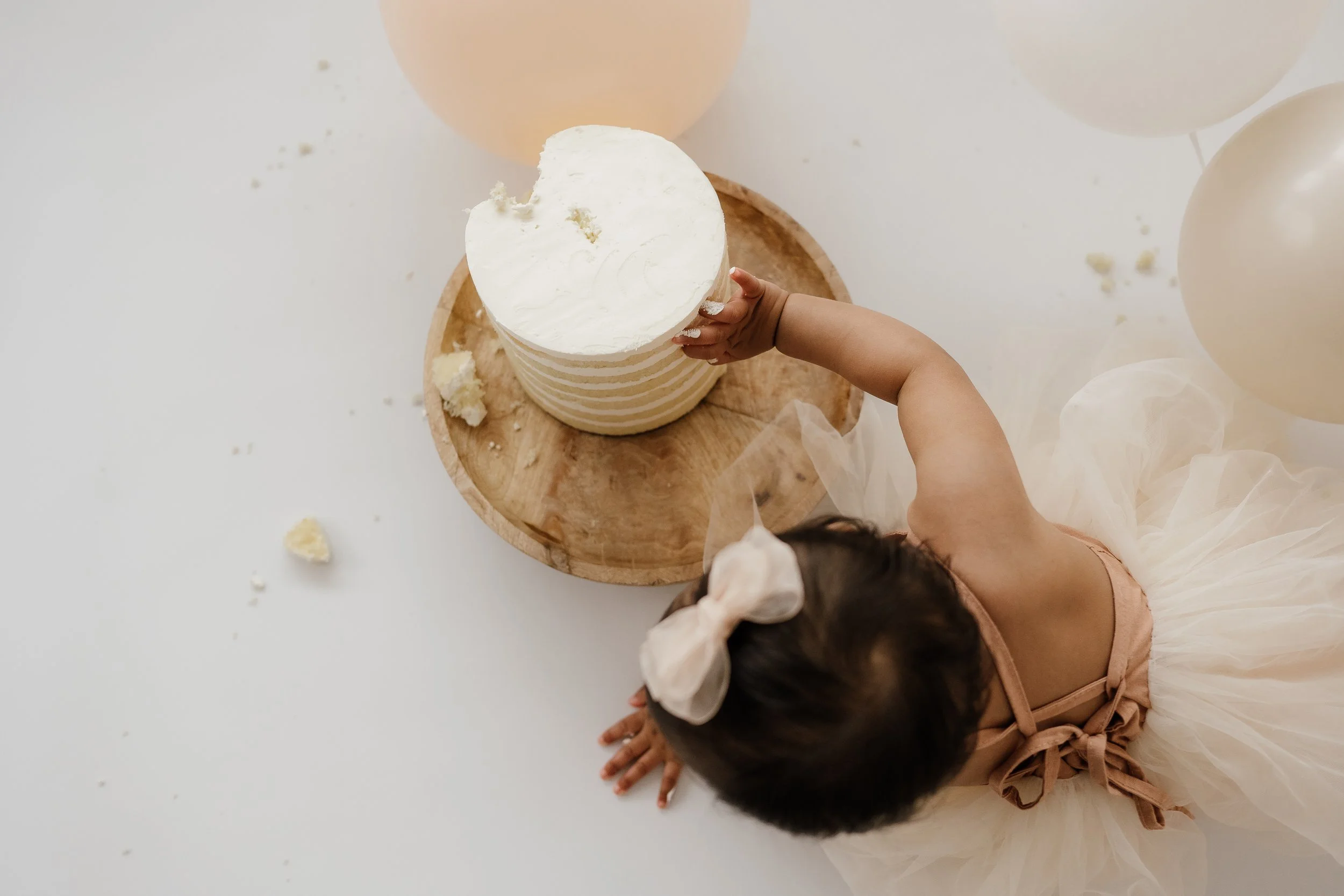 A young girl with dark hair in a light-colored dress and bow in her hair reaching into a partially eaten layered cake with white frosting, on a wooden tray, surrounded by balloons and white tulle fabric.