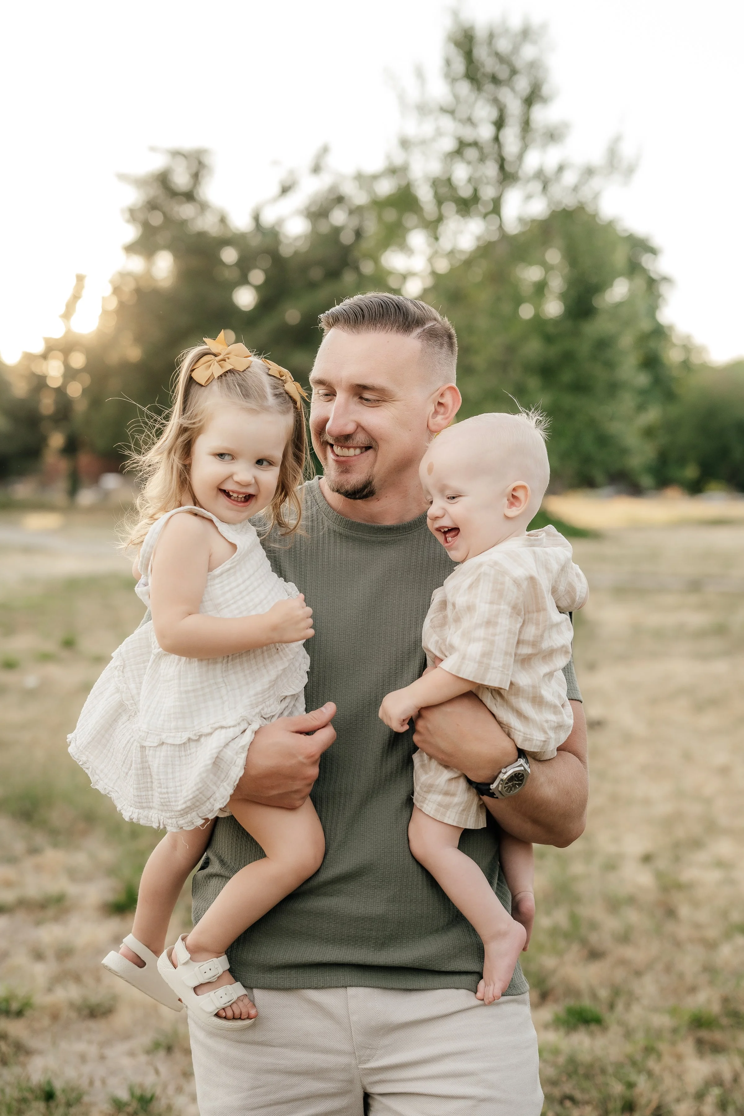 A man holding a young girl and a young boy outdoors in a park during sunset, all smiling and enjoying the moment.