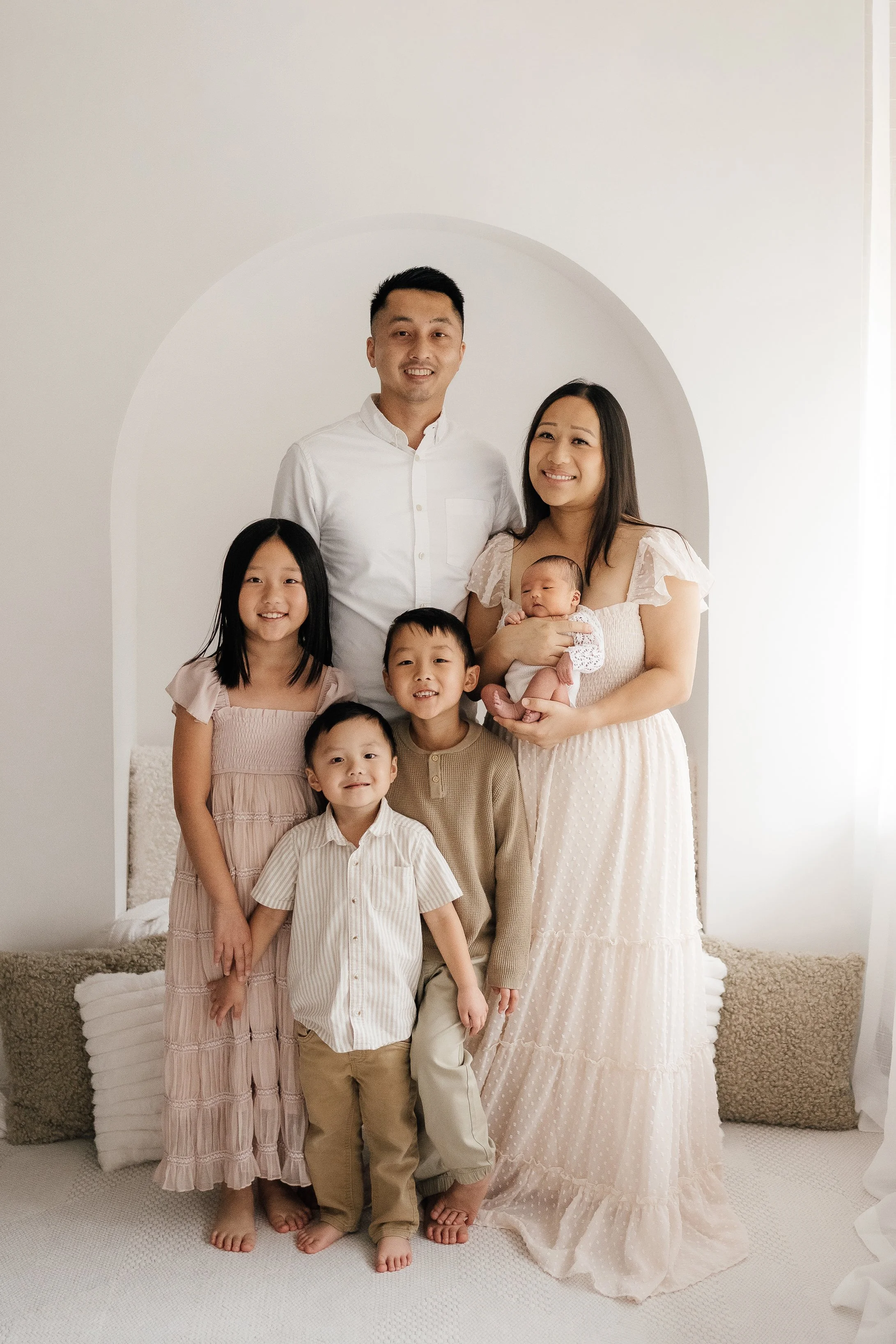 A family of six posing in front of a white wall with a rounded arch. The father and mother are standing in the back, with the mother holding a newborn. Three children are standing in front of the parents, smiling. The family appears happy and well-dr