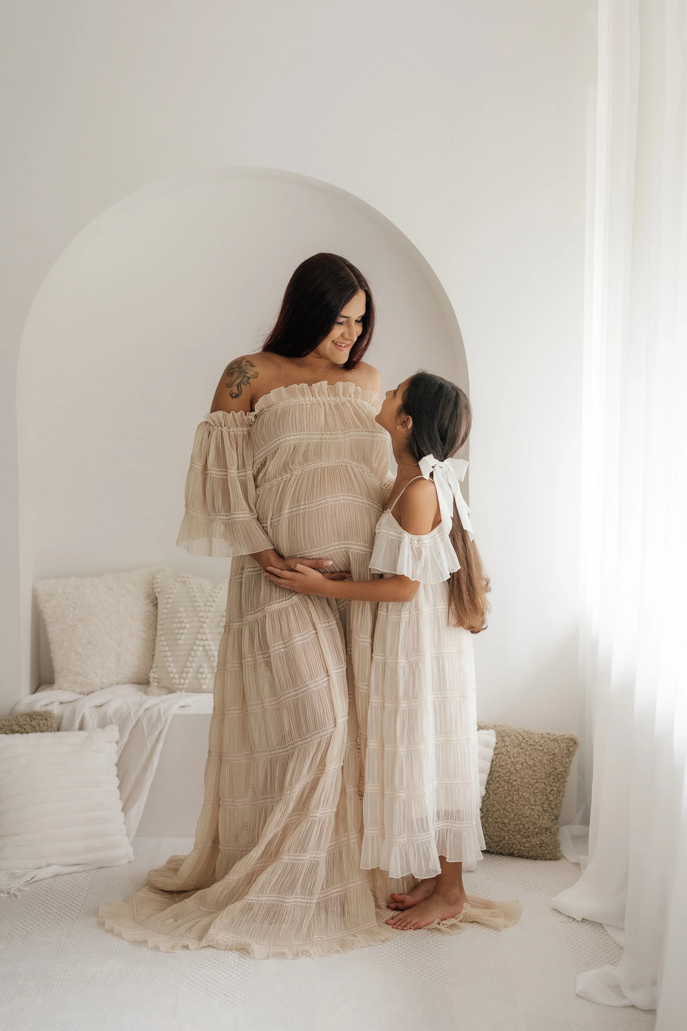 A pregnant woman and a young girl, possibly her daughter, smiling and looking at each other in a bright, white room decorated with soft cushions and sheer curtains.
