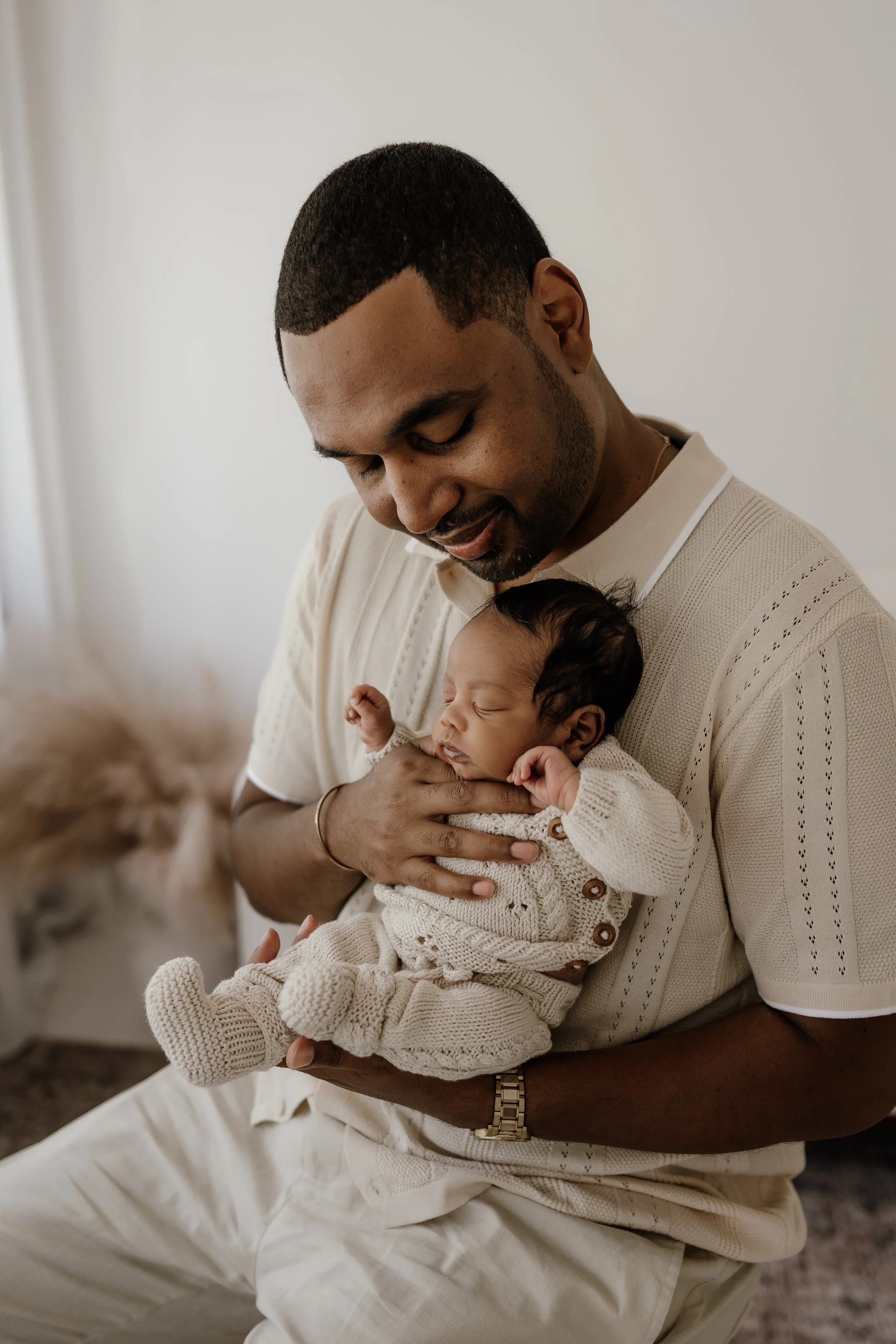An African American man holding a sleeping baby dressed in cream-colored knitted clothes, looking down with a gentle smile in a softly lit room.