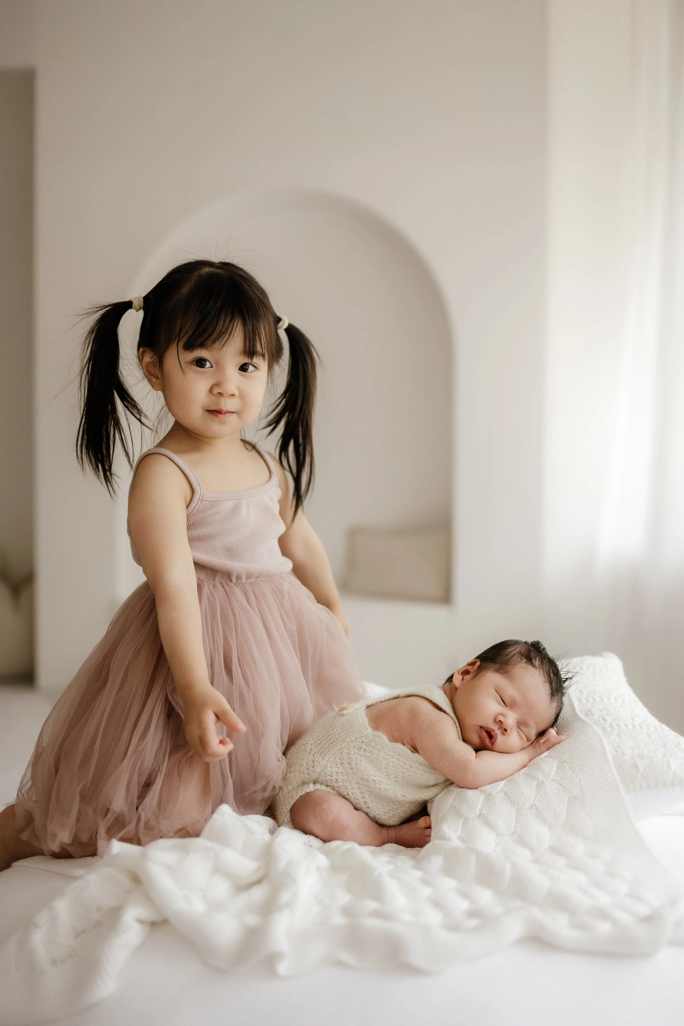 A young girl with pigtails wearing a pink dress stands next to a sleeping baby on a bed with white bedding in a softly lit room.
