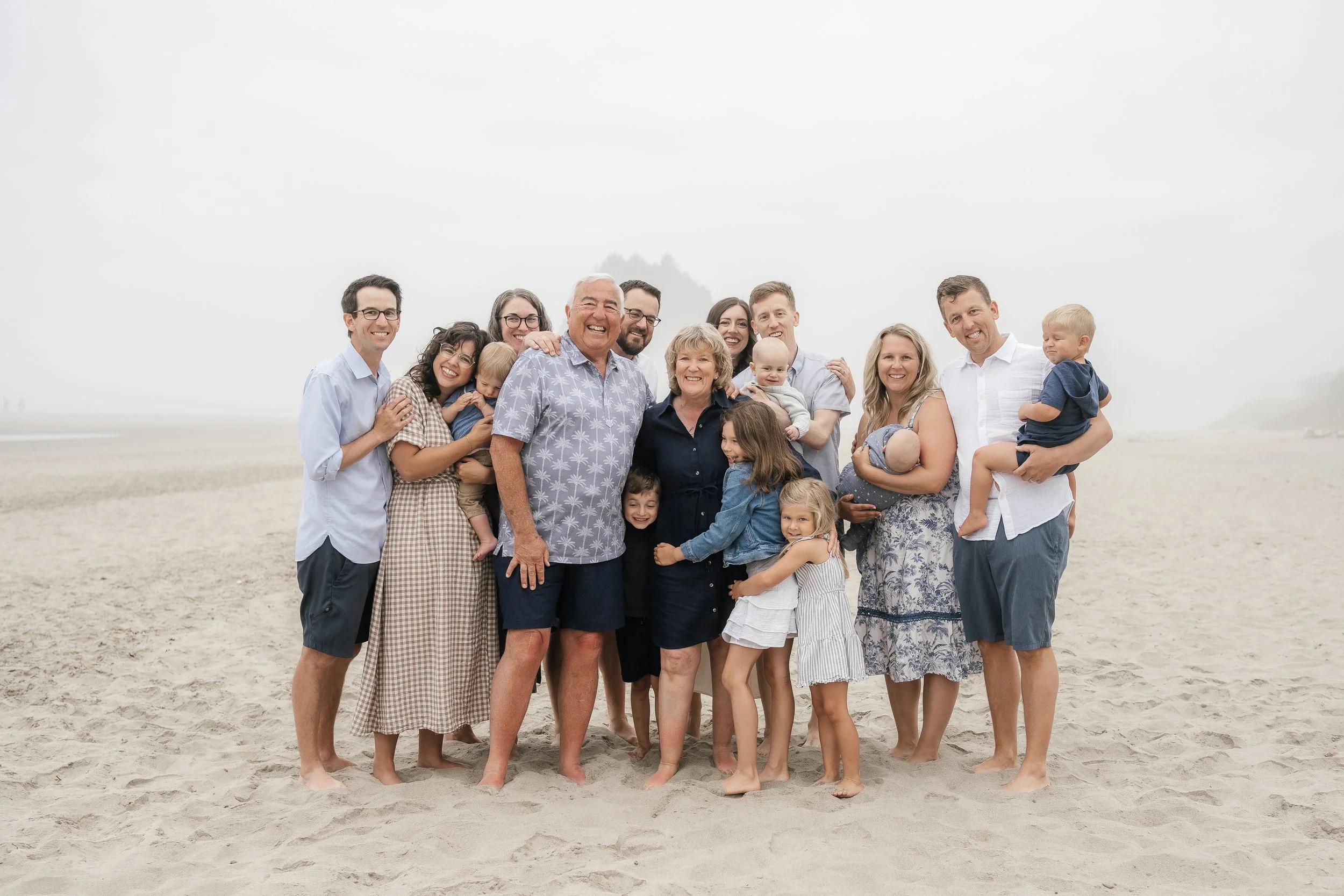 Large family group standing on sandy beach on a foggy day, smiling and hugging each other.